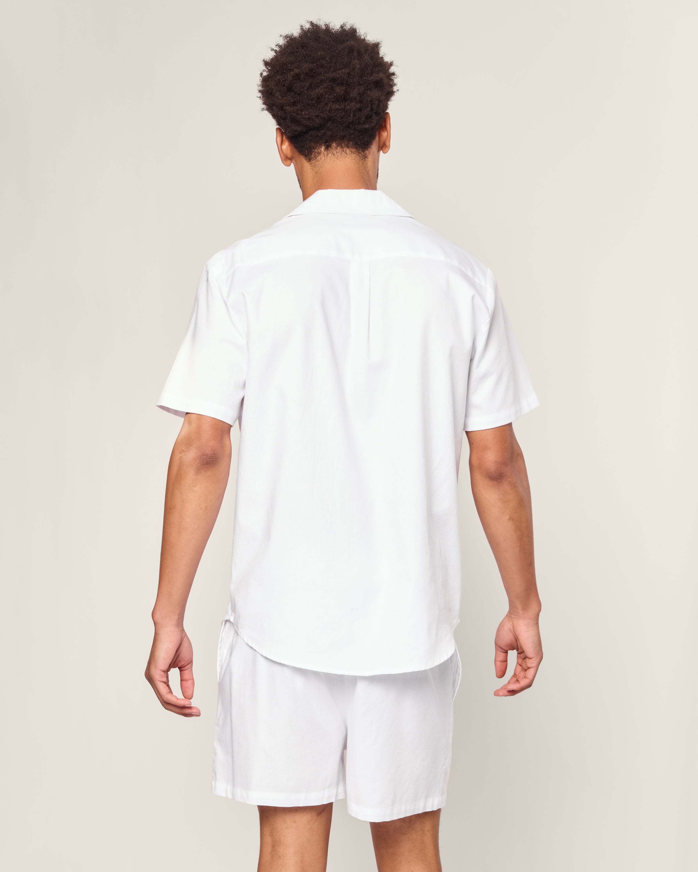 A person stands with their back to the camera, wearing the Mens Twill Pajama Short Set White with White Piping by Petite Plume. The background is a neutral beige color.