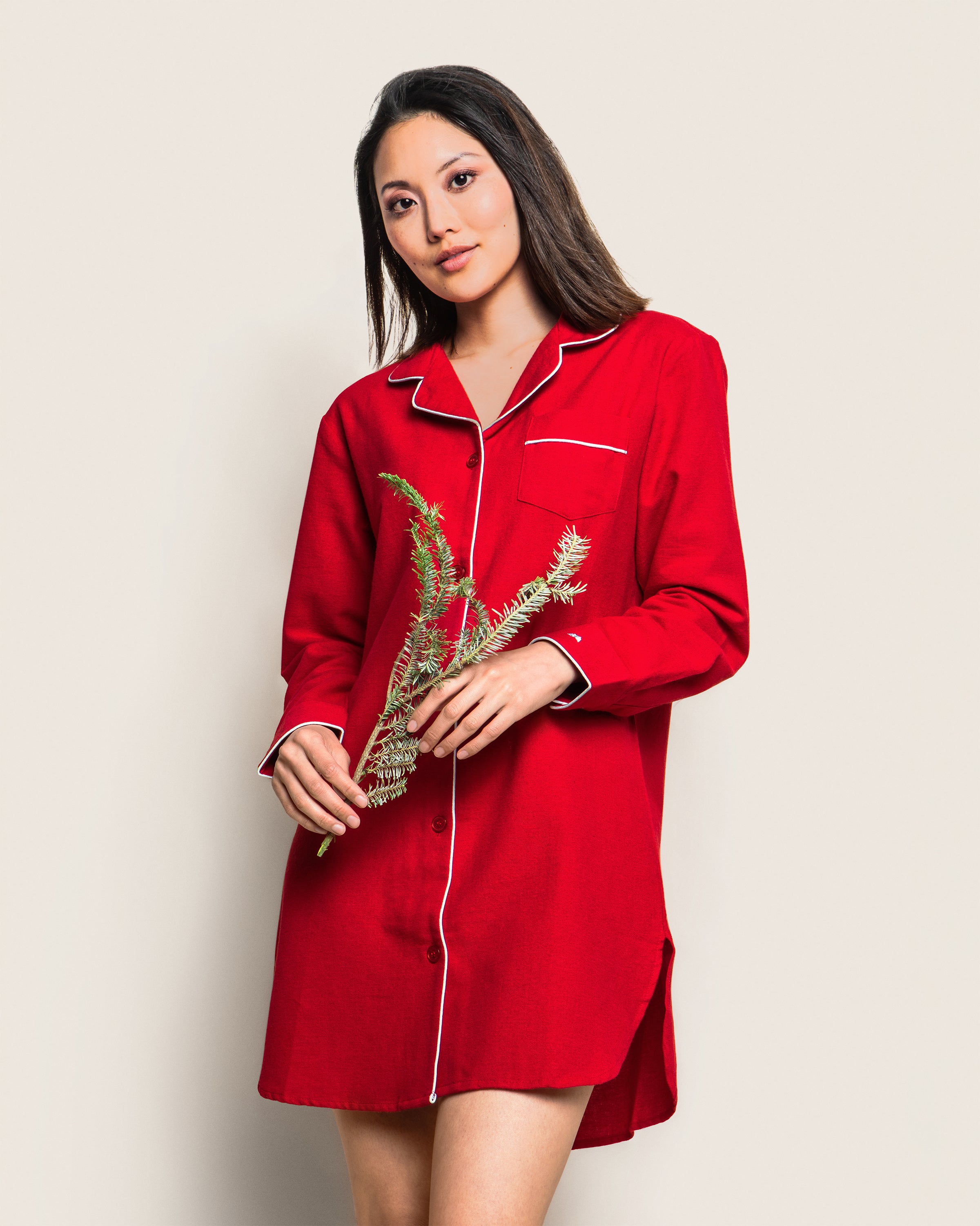 A woman wearing the luxurious Womens Flannel Nightshirt in Red by Petite Plume, featuring white trim, holds a sprig of greenery. She stands against a plain light background, looking towards the camera with a relaxed expression.