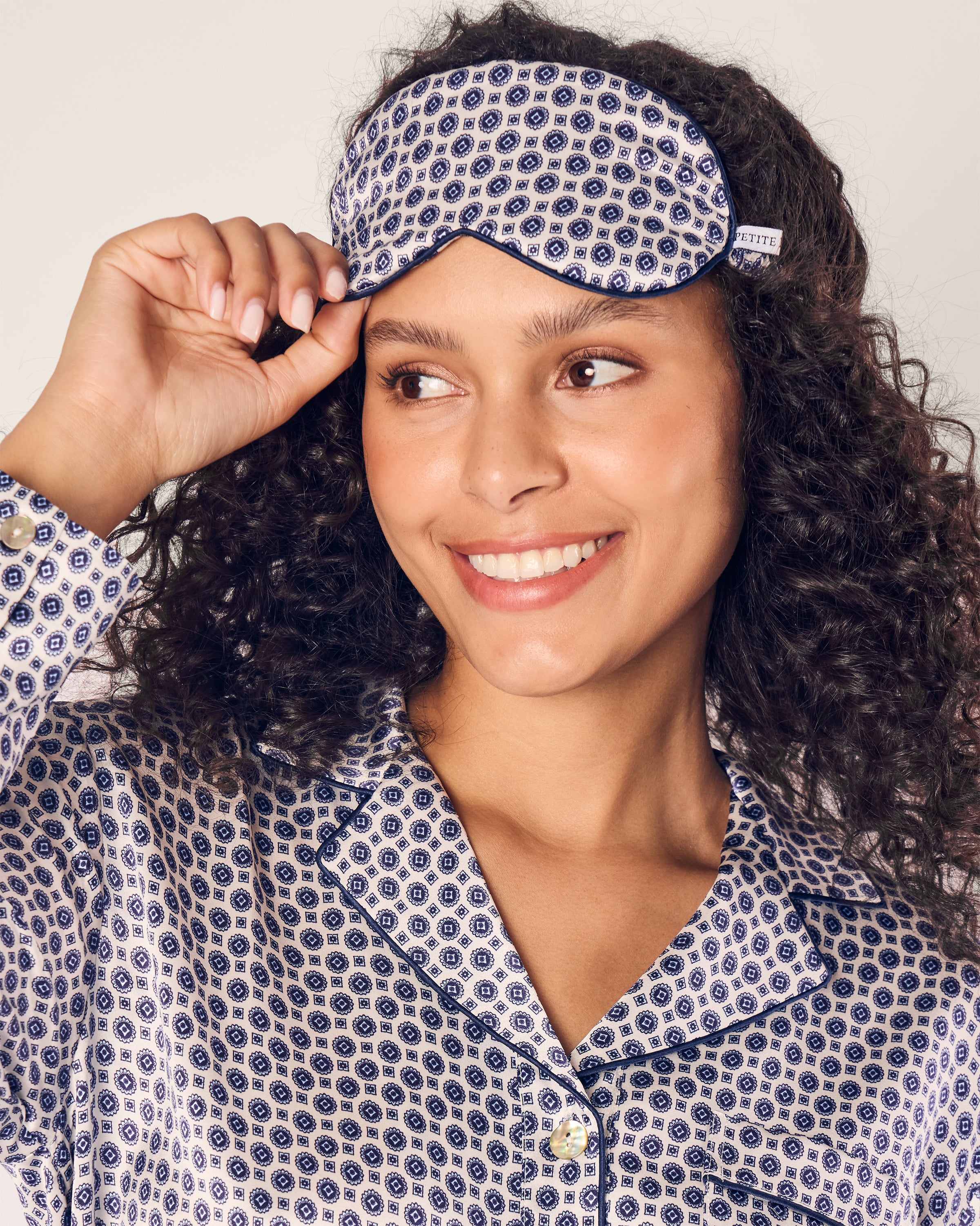 A person with curly hair smiles while adjusting a Petite Plume Adults Silk Sleep Mask in Navy Foulard on their forehead, paired with matching patterned pajamas featuring a circular design.