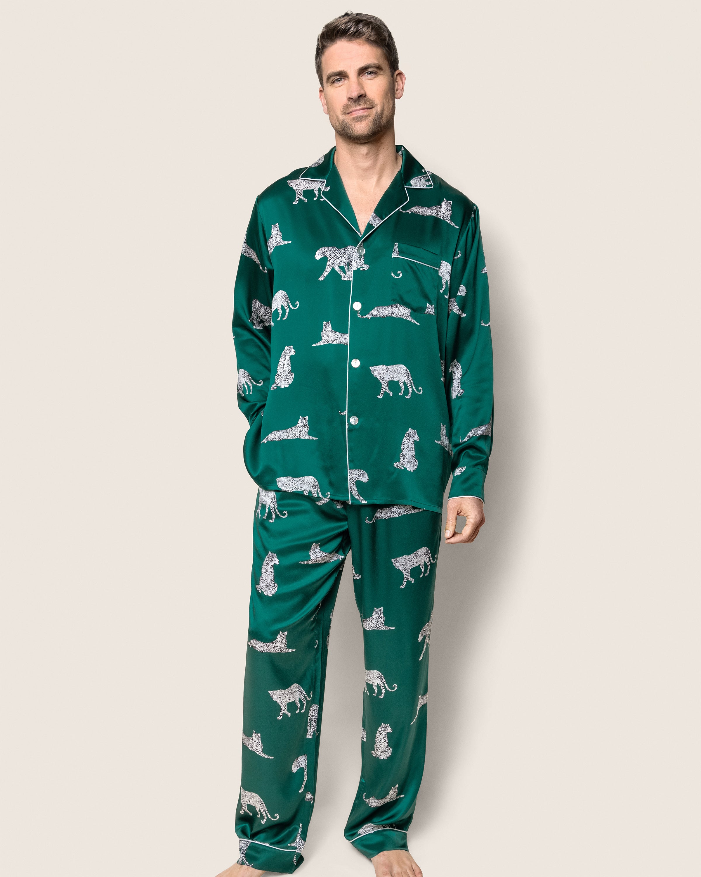 A person wearing Petite Plumes Mens Silk Pajama Set in Panthère de Luxe, a luxurious emerald green outfit with leopard print, stands against a plain background.