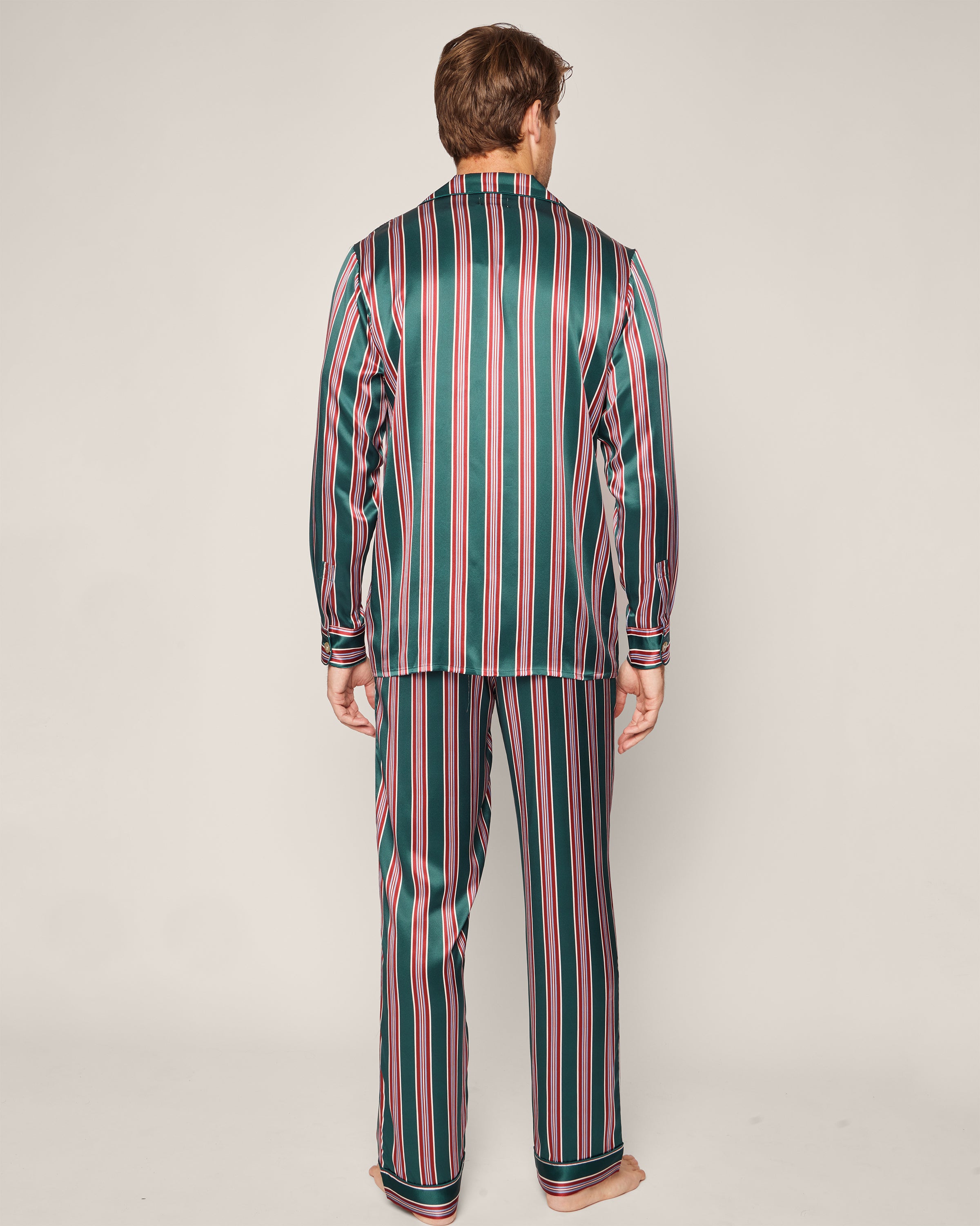 A person, facing away, wears the luxurious Petite Plume Mens Silk Pajama Set in Lancaster Stripe with temperature-regulating silk and green, red, and white vertical stripes against a plain light-colored background.
