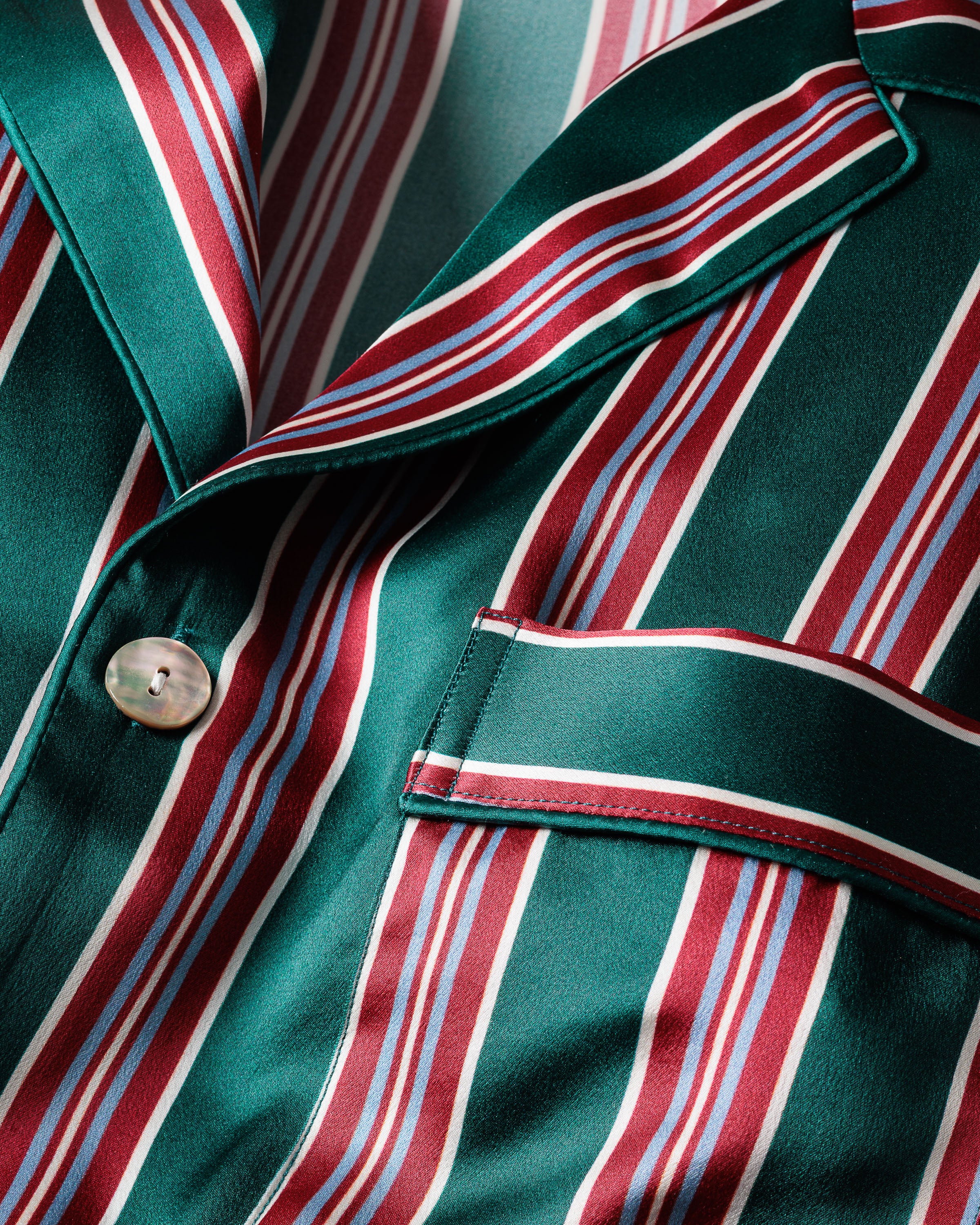 A close-up of a luxurious green silk fabric with red, blue, and white stripes from the Mens Silk Pajama Set in Lancaster Stripe by Petite Plume. The smooth, shiny texture features a button and pocket, suggesting it belongs to temperature-regulating sleepwear.