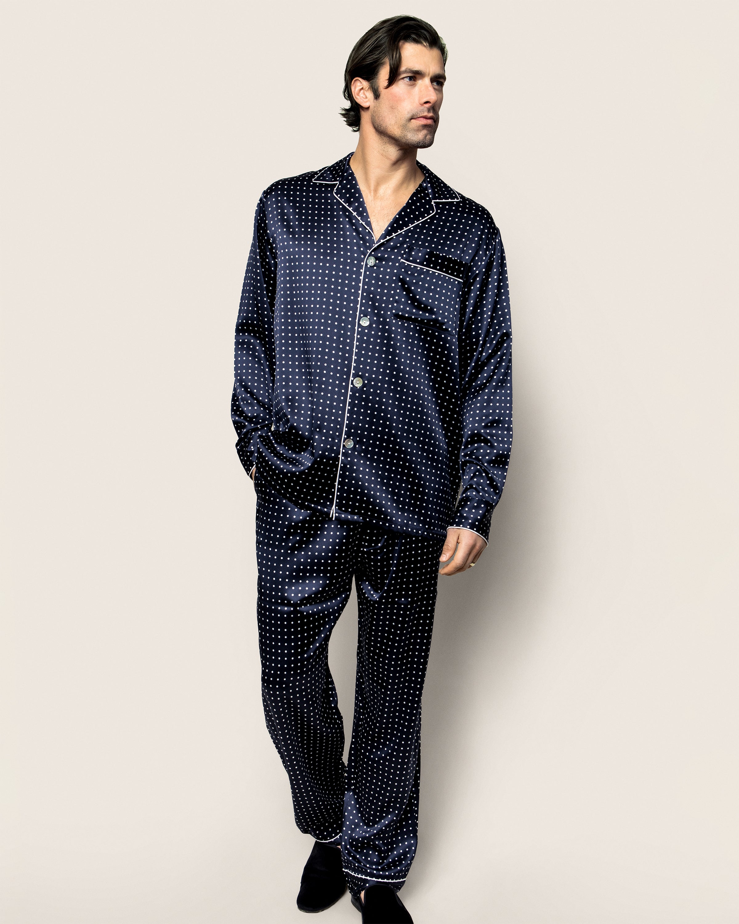 A man in the Petite Plume Mens Silk Pajama Set in Navy Polka Dot, featuring mulberry silk with white polka dots, stands against a plain beige background. He wears a button-up shirt and matching pants, has dark hair, and looks to his left.