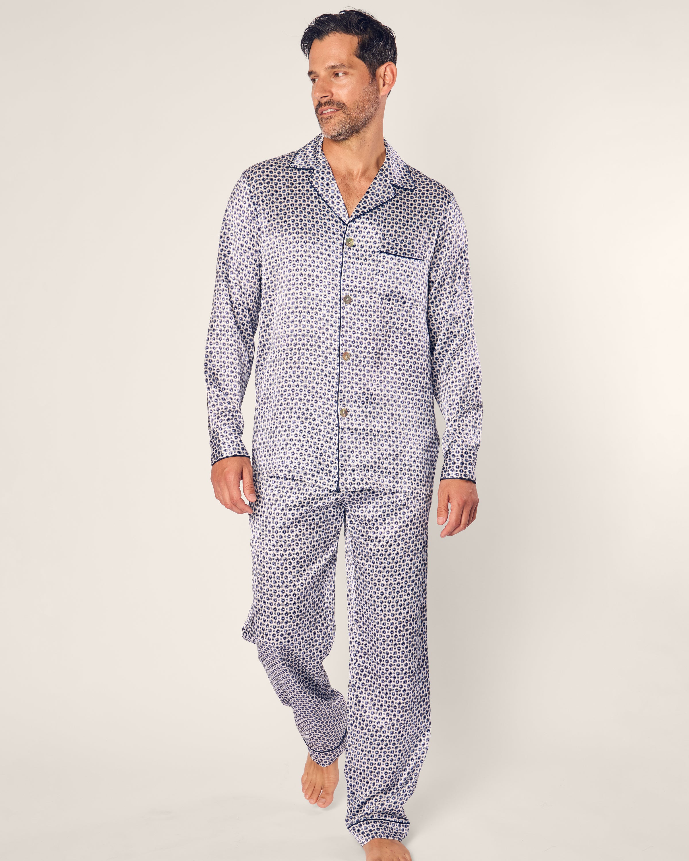 A man walks barefoot on a light background wearing Petite Plumes Mens Silk Pajama Set in Navy Foulard. The luxurious mulberry silk set features a button-up shirt and pants with a small dark geometric pattern, giving him a relaxed expression.
