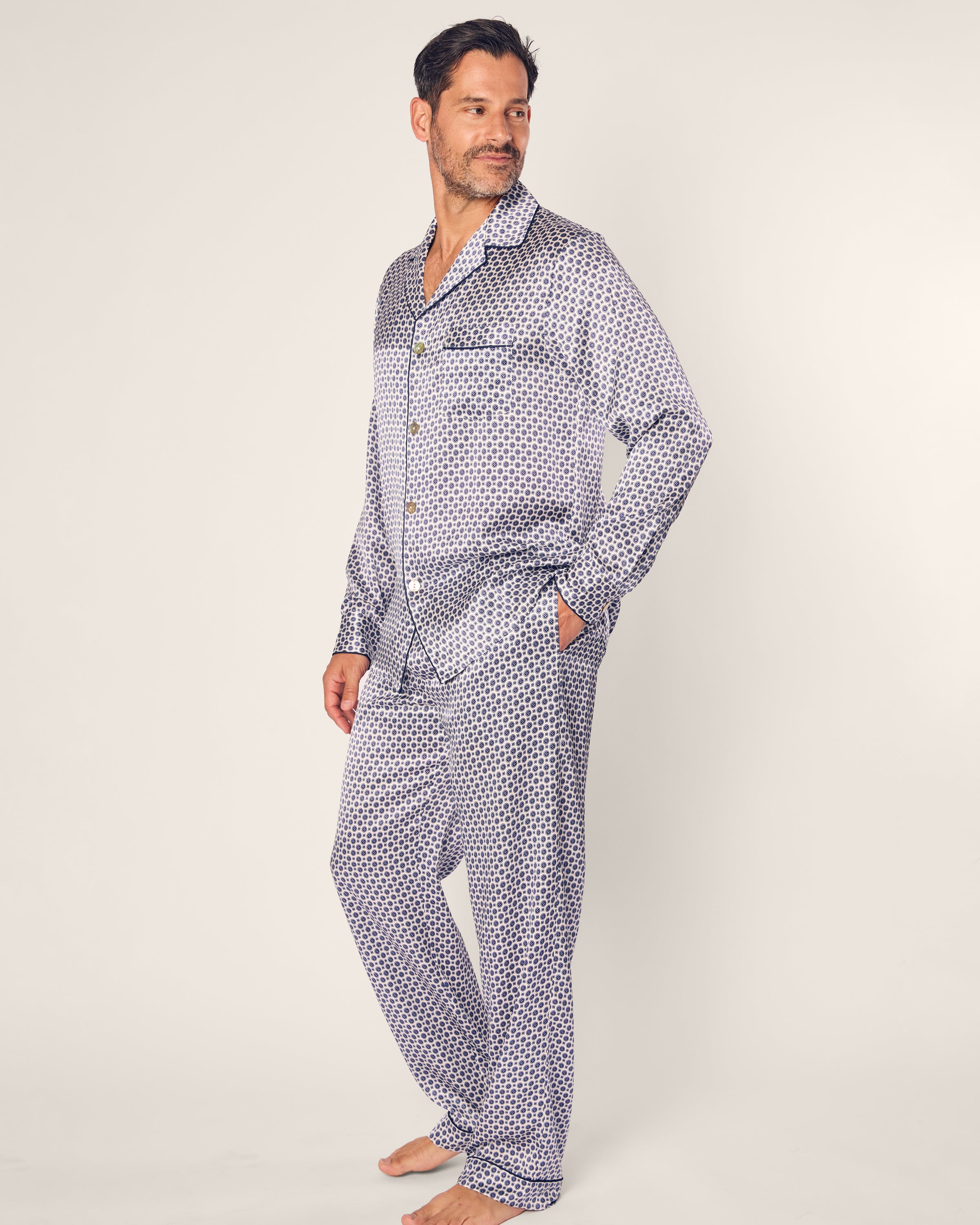 A man stands barefoot in a luxurious Petite Plume Mens Silk Pajama Set in Navy Foulard, featuring a small geometric pattern. With his hands in his pockets and looking left with a slight smile, he is set against a plain white background.