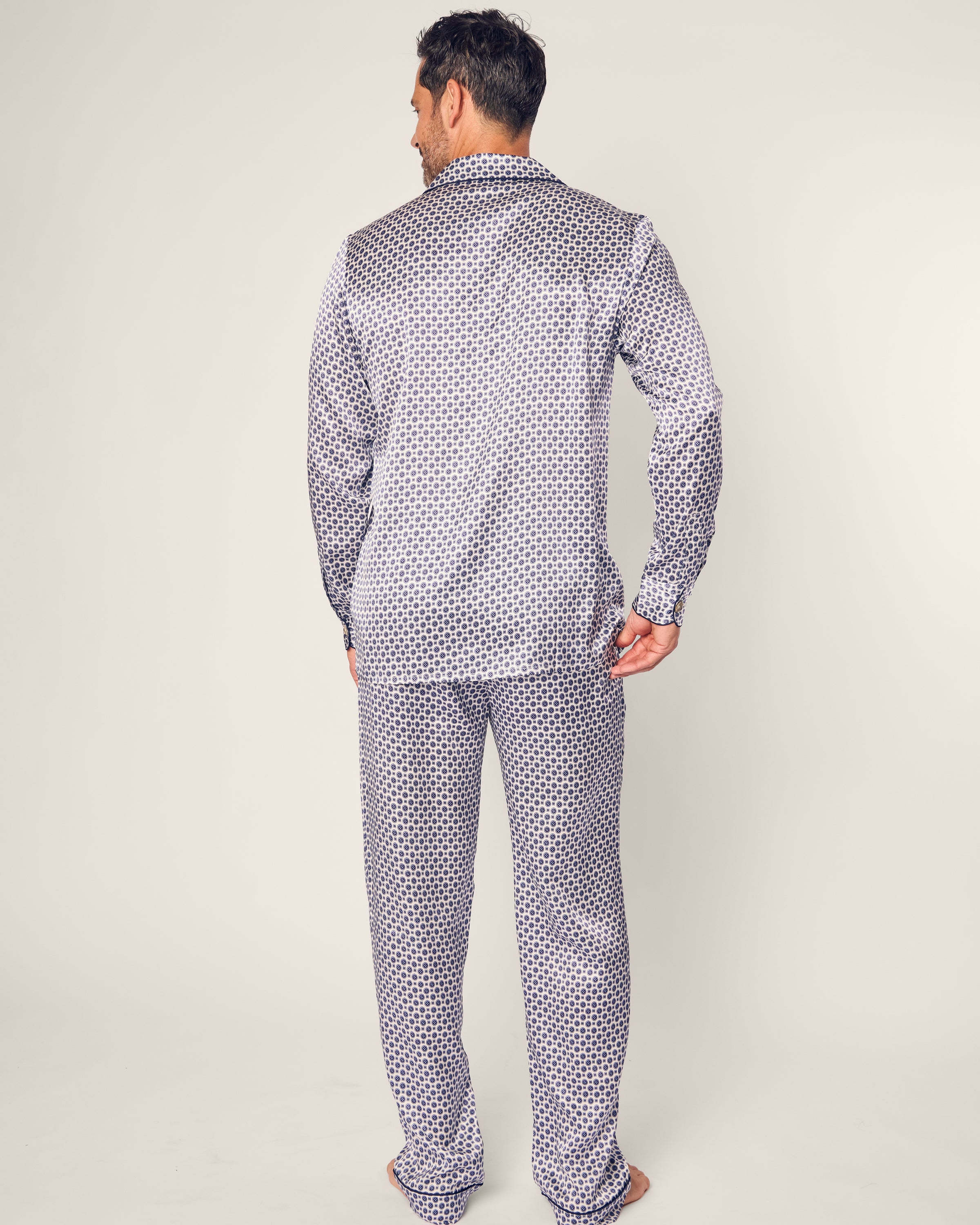 A man stands barefoot against a plain backdrop, wearing the Mens Silk Pajama Set in Navy Foulard by Petite Plume, featuring a geometric pattern. He faces away from the camera, embodying an elegant take on luxury sleepwear.