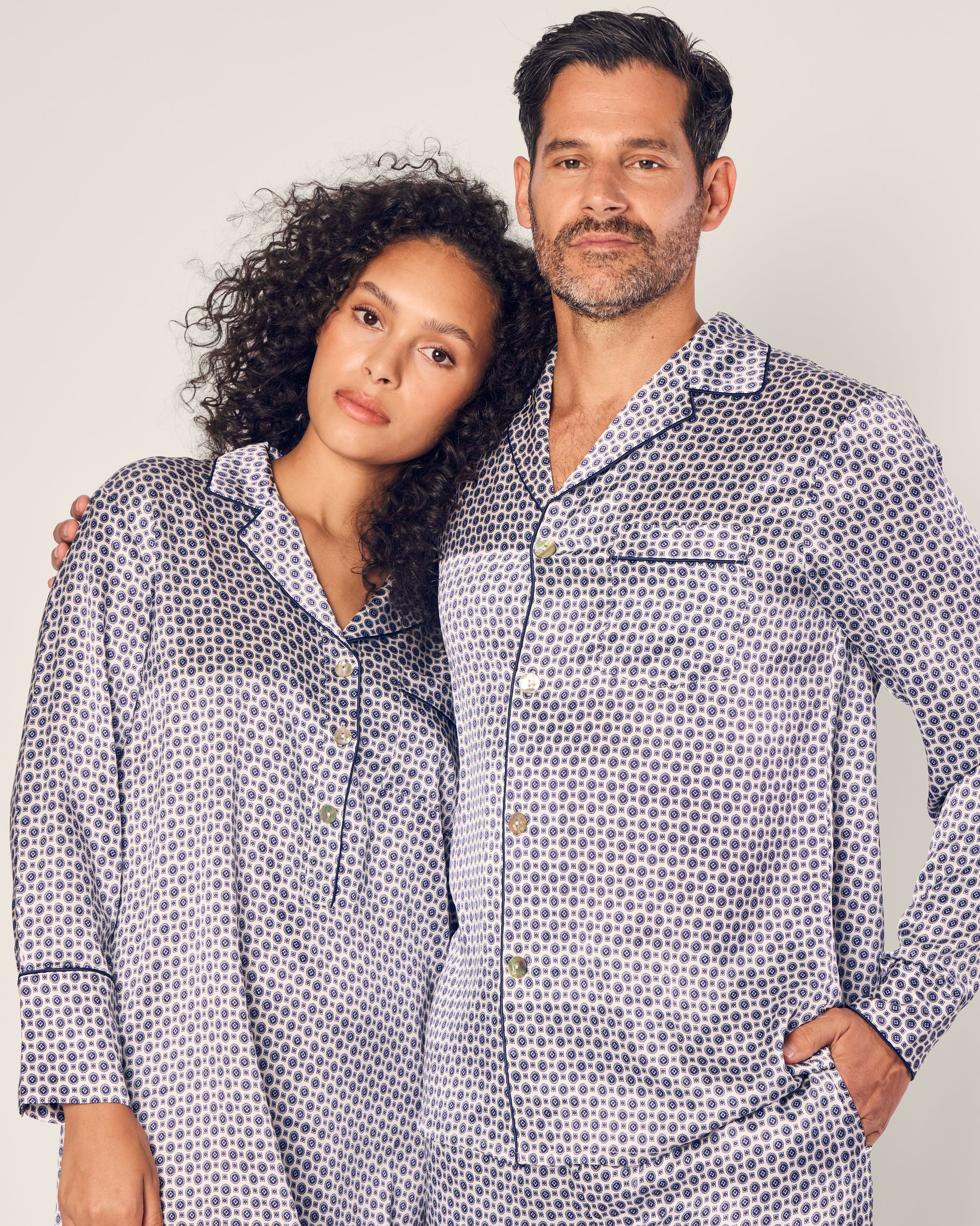A relaxed couple in matching Petite Plume pajamas, made of luxurious mulberry silk with dark circular patterns, stands together. The pajamas worn by the person on the left complement their curly hair, while the person on the right displays short hair and stubble.