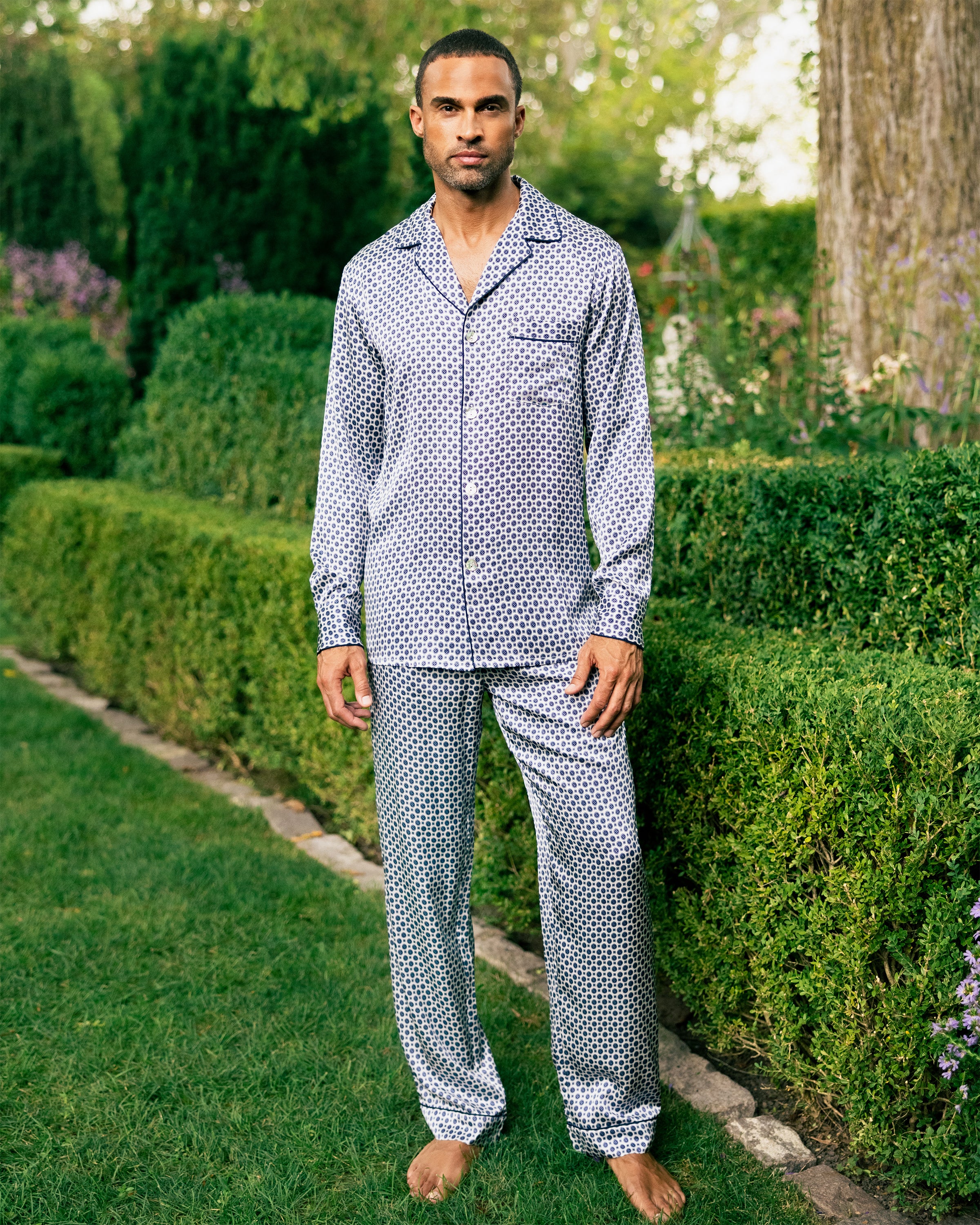 A man stands barefoot on grass, wearing the Petite Plume Mens Silk Pajama Set in Navy Foulard. He is near a trimmed hedge with tall trees and various plants visible in the background on a sunny day.