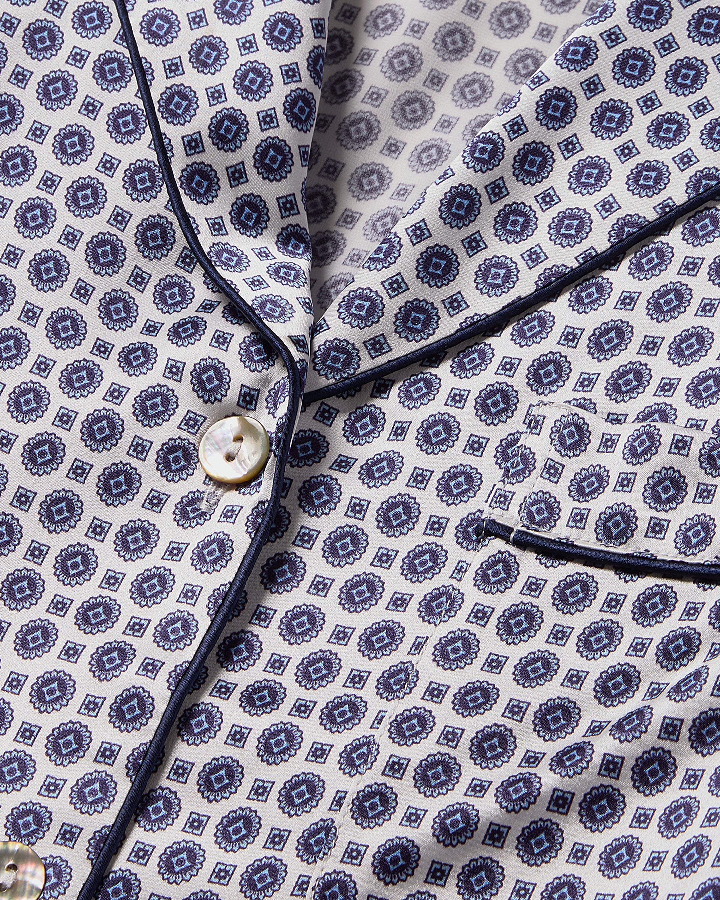 Close-up of the Petite Plume Mens Silk Pajama Set in Navy Foulard, showcasing a geometric blue pattern on white silk with button closure and piped trim along the collar and pocket edge for a classic, elegant design.