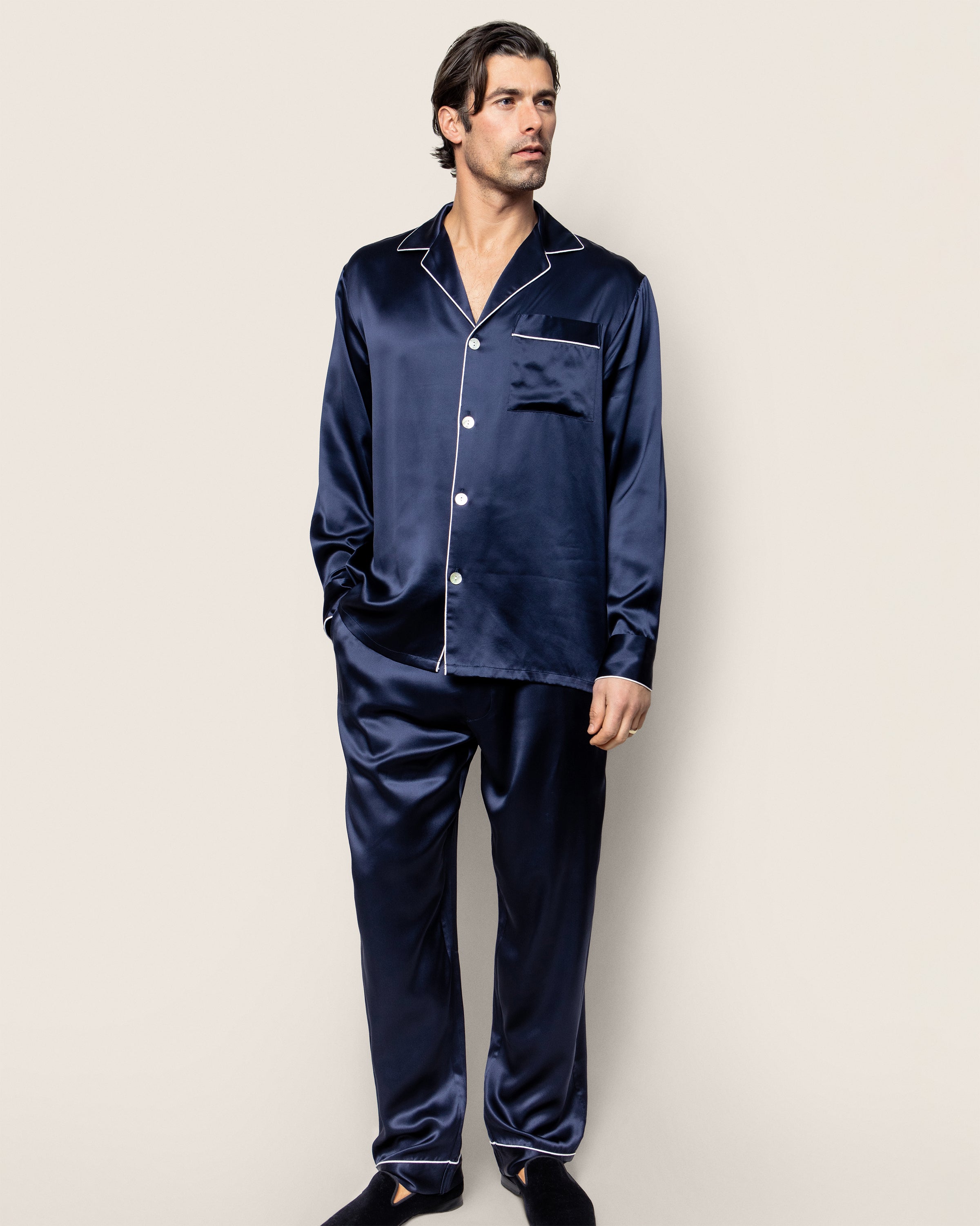 A person is wearing a Petite Plume Mens Silk Pajama Set in Navy, adorned with white piping. This luxury sleepwear includes a button-up top with a chest pocket and matching pants, crafted from machine-washable silk, set against a plain light-colored background.