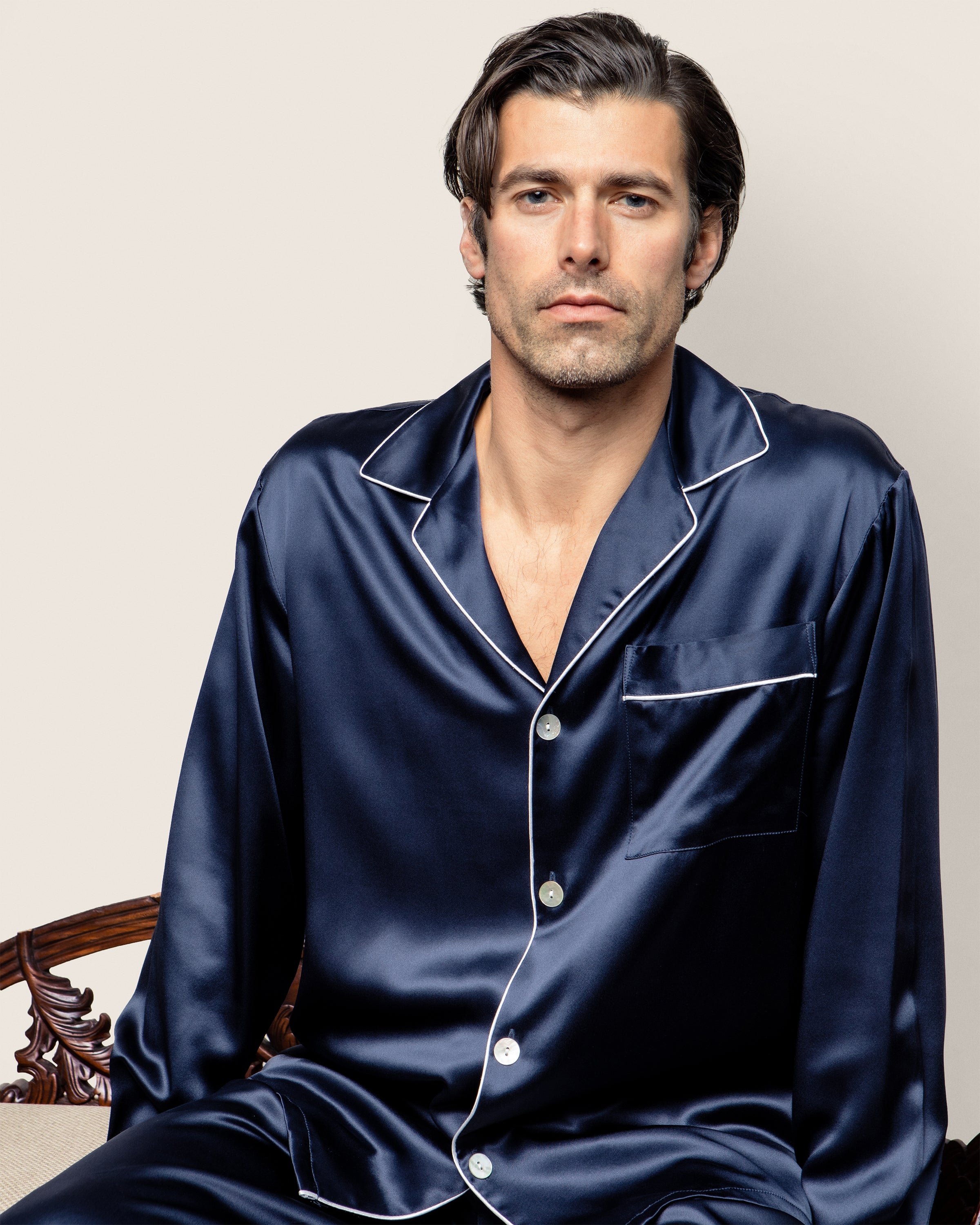 A person with short dark hair sits on a patterned wooden chair against a plain light background, dressed in the Mens Silk Pajama Set in Navy by Petite Plume—a luxurious and elegant set featuring white piping details that is also machine-washable.