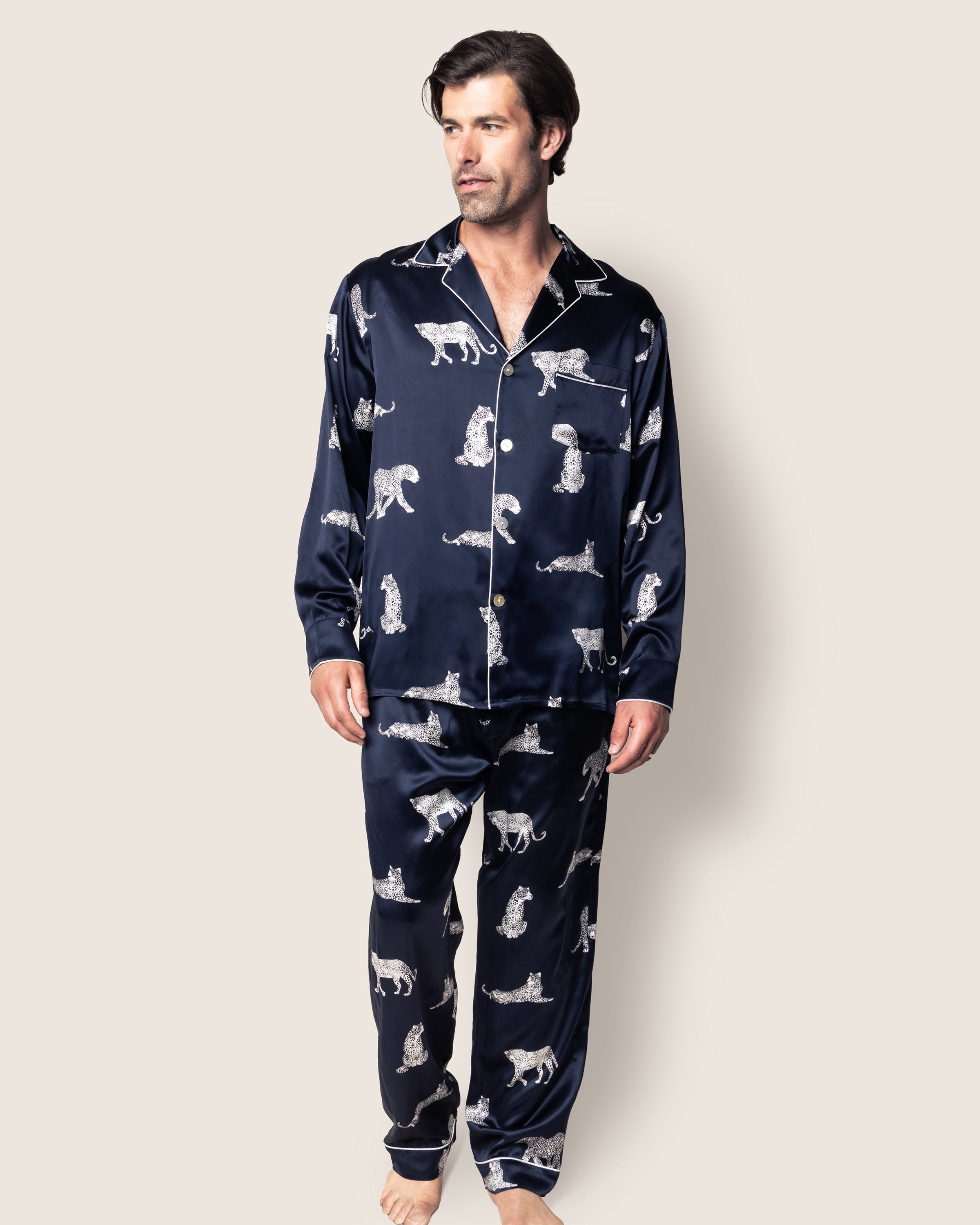 A person models the Mens Silk Pajama Set in Panthère de Nuit by Petite Plume, embodying classic Hollywood style. The dark blue top sports a collar, buttons, and white animal prints, while the loose pants ensure comfort. The luxurious hypoallergenic silk echoes elegance against a plain background with bare feet.