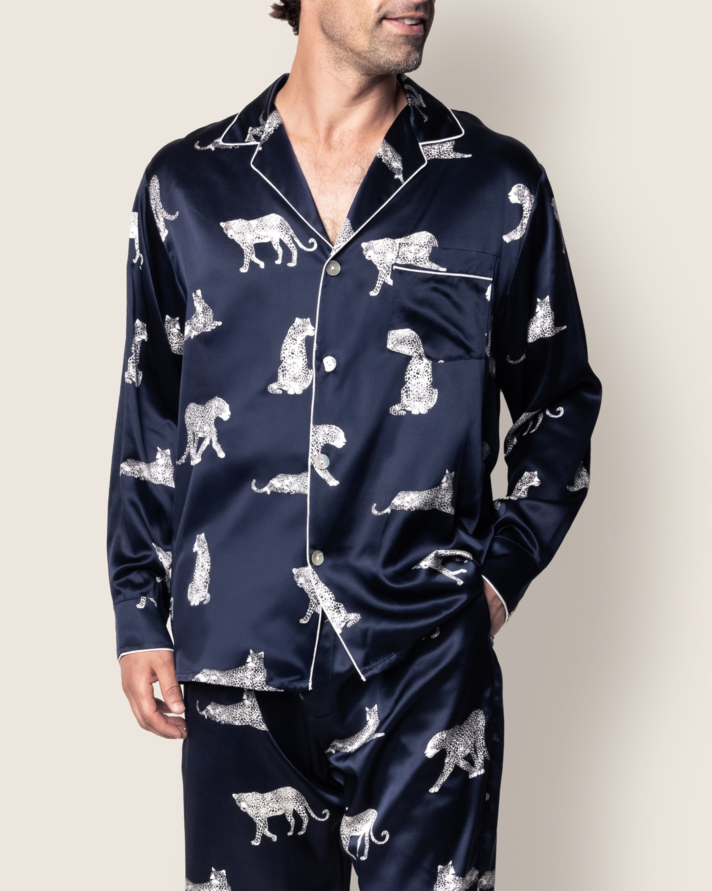 A person wearing the Mens Silk Pajama Set in Panthère de Nuit by Petite Plume exudes classic Hollywood elegance. The dark blue satin set, adorned with leopard illustrations, features a top with a front pocket and white piping, as they stand relaxed with hands partly in their pant pockets.