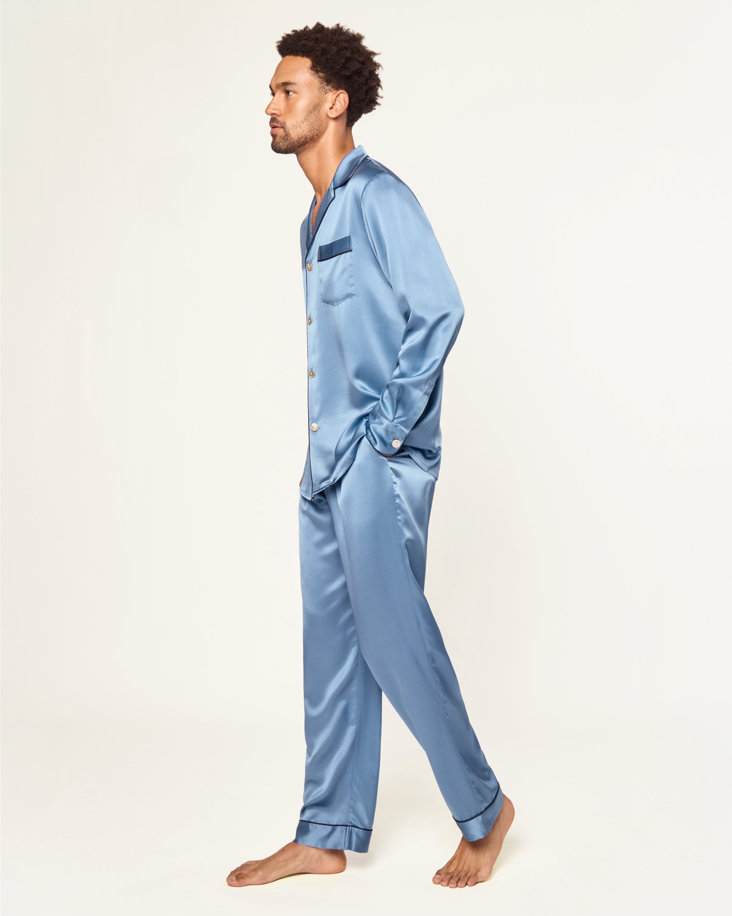 The Men's Silk Pajama Set in Mystique Blue features a button-up long-sleeve top with a chest pocket and matching elastic-waist pants, both trimmed with dark piping.