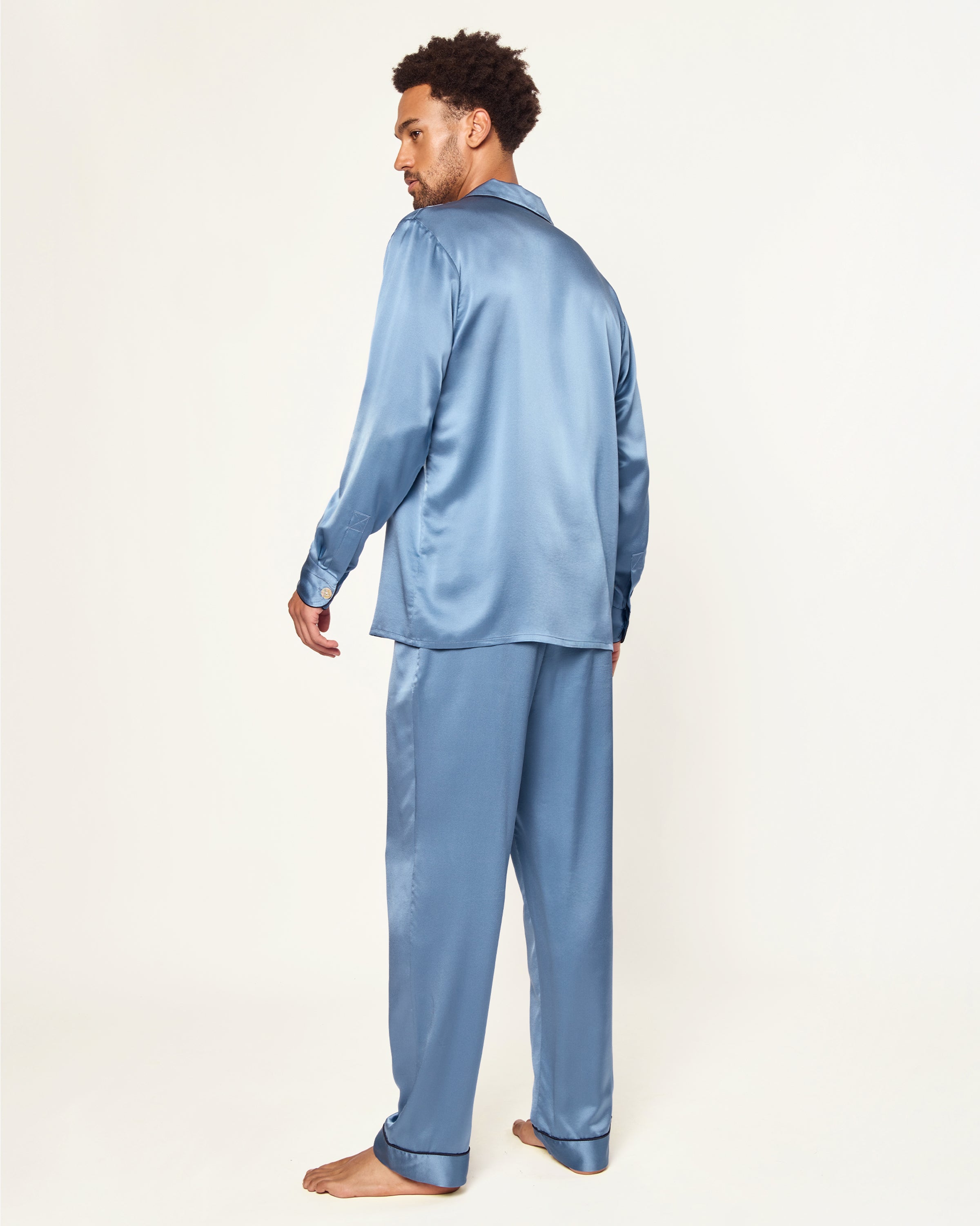 The Men's Silk Pajama Set in Mystique Blue features a button-up long-sleeve top with a chest pocket and matching elastic-waist pants, both trimmed with dark piping.