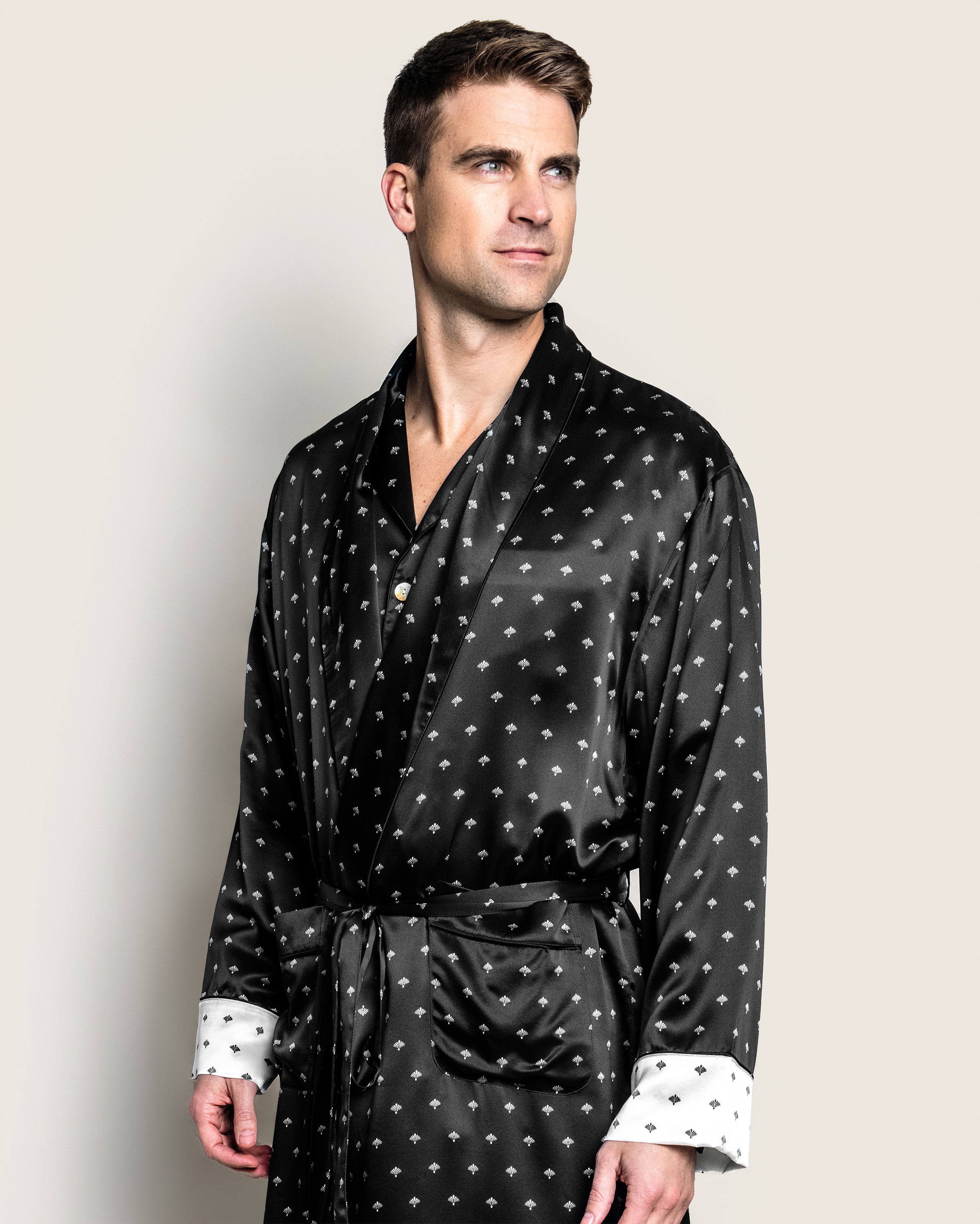A man wearing Petite Plumes Mens Silk Long Robe in Black Art Nouveau, featuring white cuffs and tied at the waist, stands out against a plain, light backdrop as he gazes off to the side.