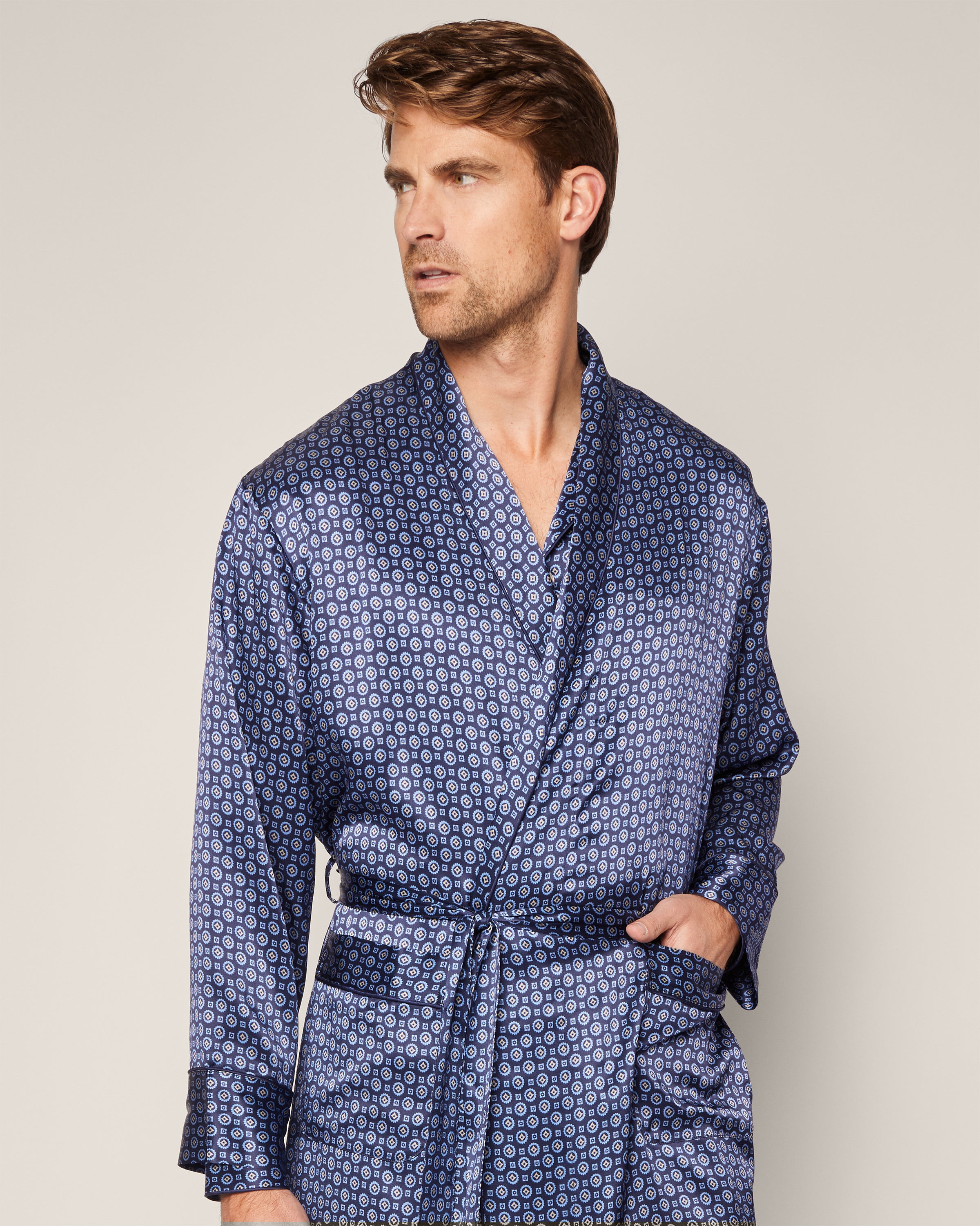 A man stands against a plain backdrop, wearing Petite Plumes Mens Silk Robe in Midnight Foulard, crafted from luxurious mulberry silk. Featuring red and white accents and an elegant circular design, the breathable sleepwear showcases his left hand casually tucked into a pocket as he gazes sideways.