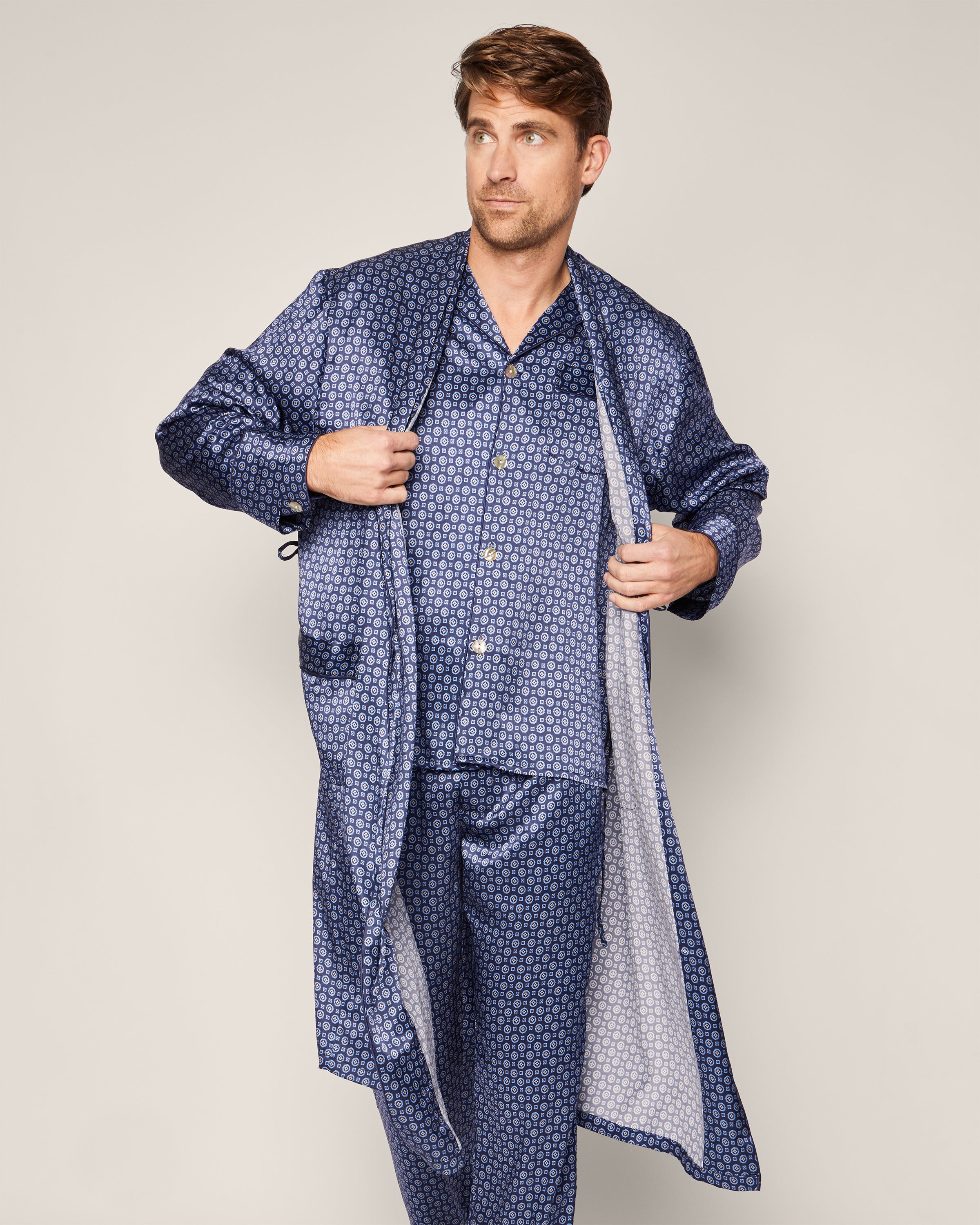 Dressed in a blue patterned pajama set and the Mens Silk Robe in Midnight Foulard by Petite Plume, a person stands against a plain background, adjusting the contrasting-lined silky robe. The luxurious ensemble complements their thoughtful gaze off to the side.