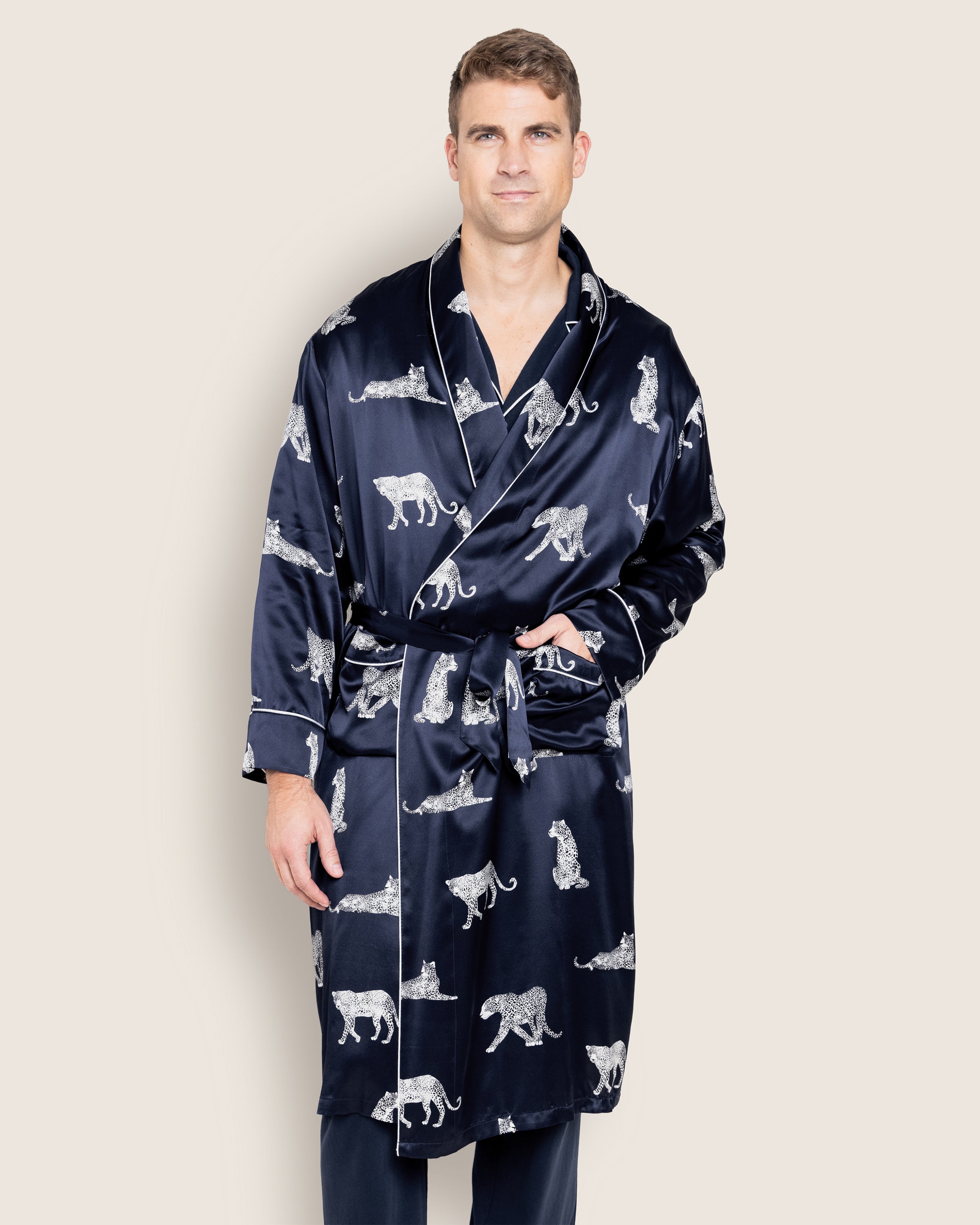 A person in a Petite Plume Mens Silk Long Robe in Panthère de Luxe, featuring white animal motifs on navy blue mulberry silk, stands against a light background. The sleepwear has an elegantly tied waist belt, exuding comfort and sophistication.