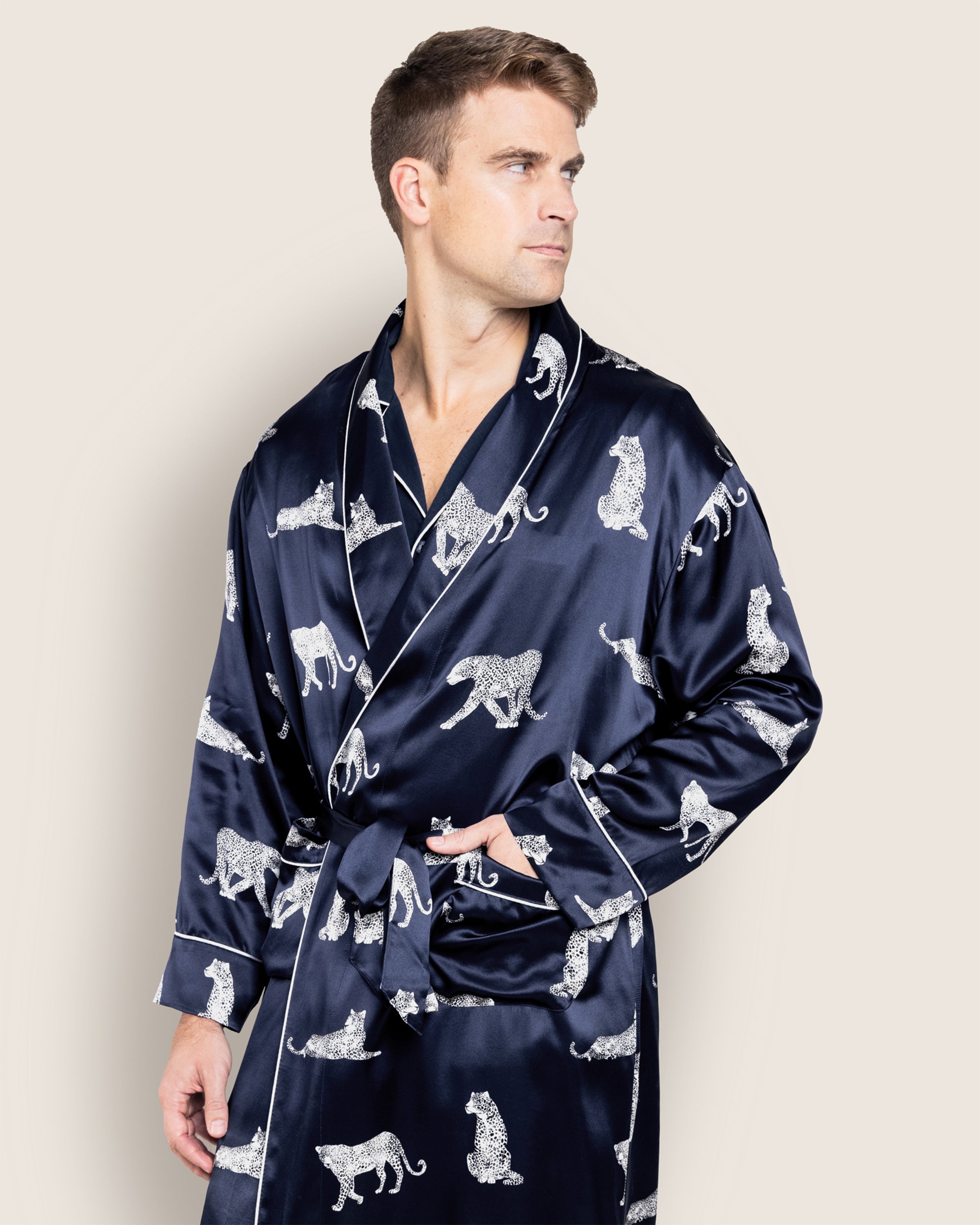 A man in a long blue silk robe, featuring the Panthère de Nuit pattern by Petite Plume, stands against a plain background. Looking to the side with his hand in a pocket, he highlights the sleepwears pattern and texture.