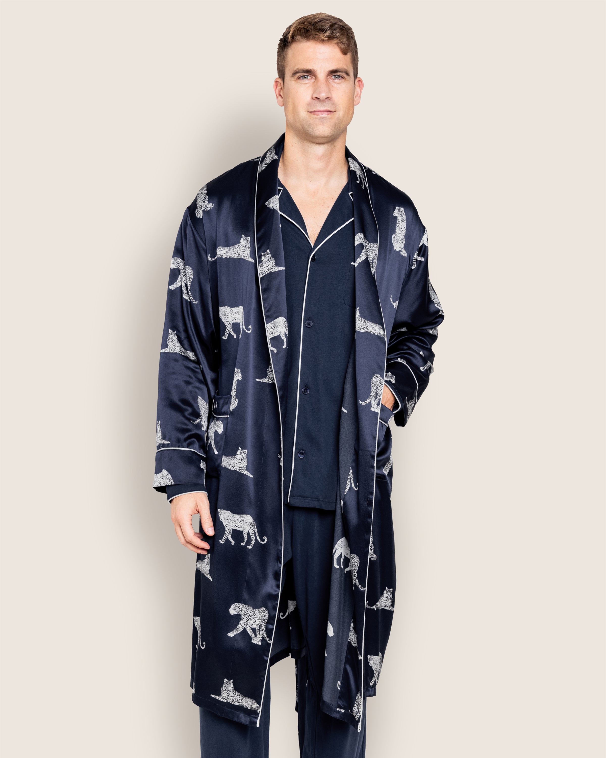 Against a plain backdrop, a person dons Petite Plumes Mens Silk Long Robe in Panthère de Nuit over dark pajamas, with one hand in a pocket. Crafted from luxurious Mulberry silk, this robe exudes elegance and comfort for serene nights.