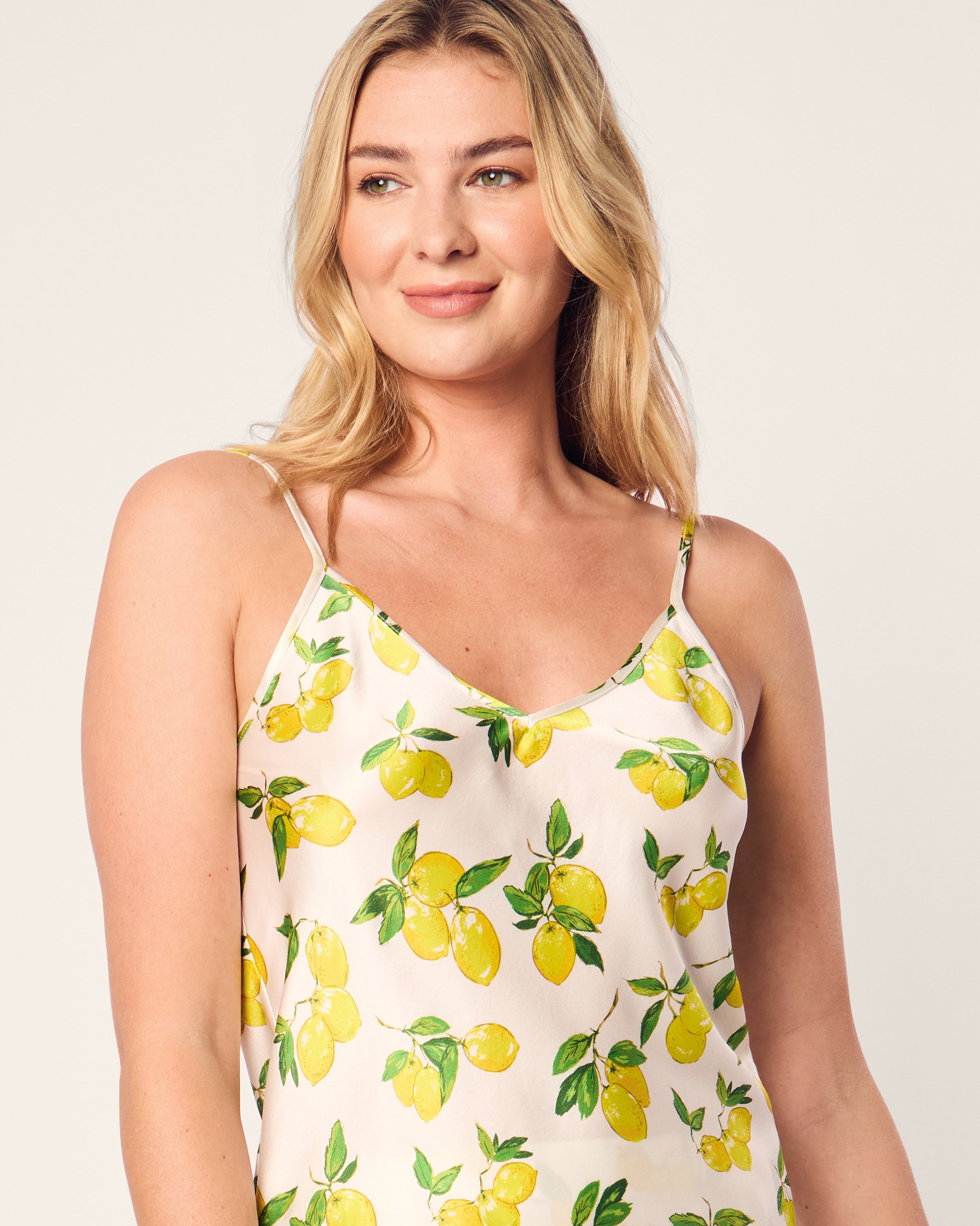 A person with long blonde hair wears the Womens Silk Short Set in Amalfi Lemons by Petite Plume, featuring a sleeveless white top with yellow lemons and green leaves, reminiscent of luxury sleepwear, against a plain, neutral background.
