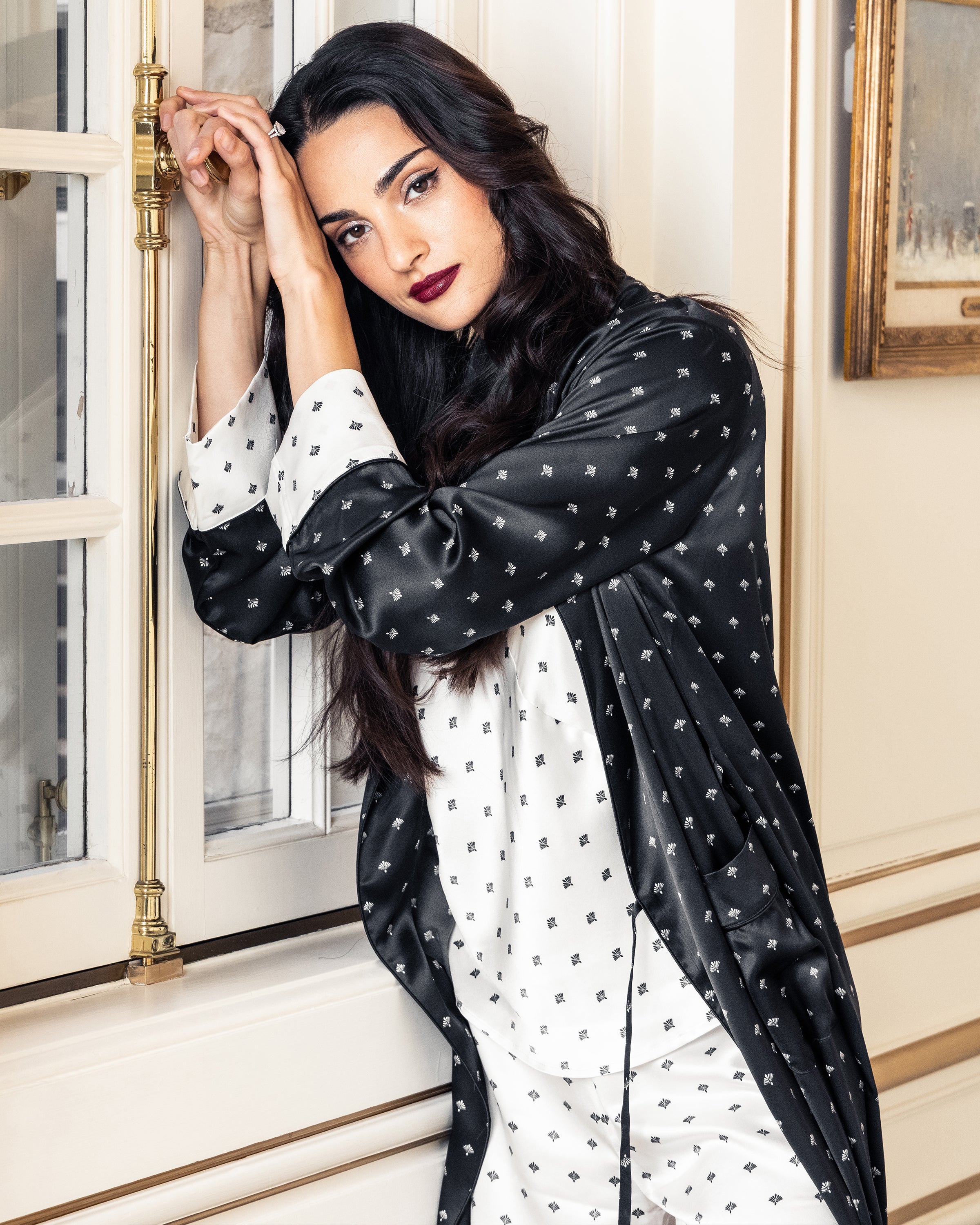 A woman with long dark hair and bold lipstick stands by a window in an elegantly decorated room, wearing Petite Plumes Womens Silk Cami Short Set in White Art Nouveau, reminiscent of an Art Nouveau print. She gazes thoughtfully, leaning against the wall.