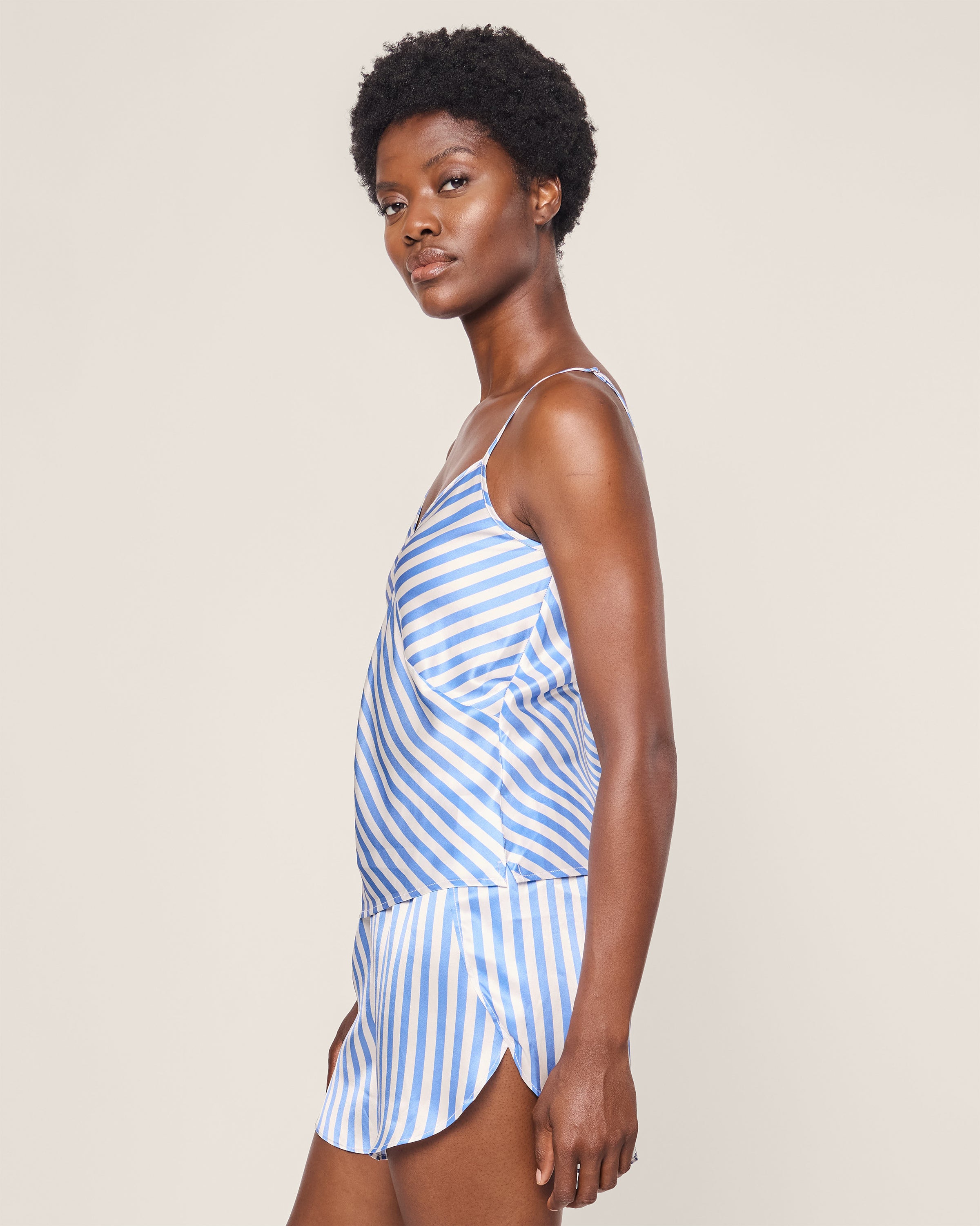 Someone wears the Womens Silk Cami Short Set in Azure Stripe by Petite Plume, shown from a side view. The plain light gray background accentuates the elegance of their outfit.