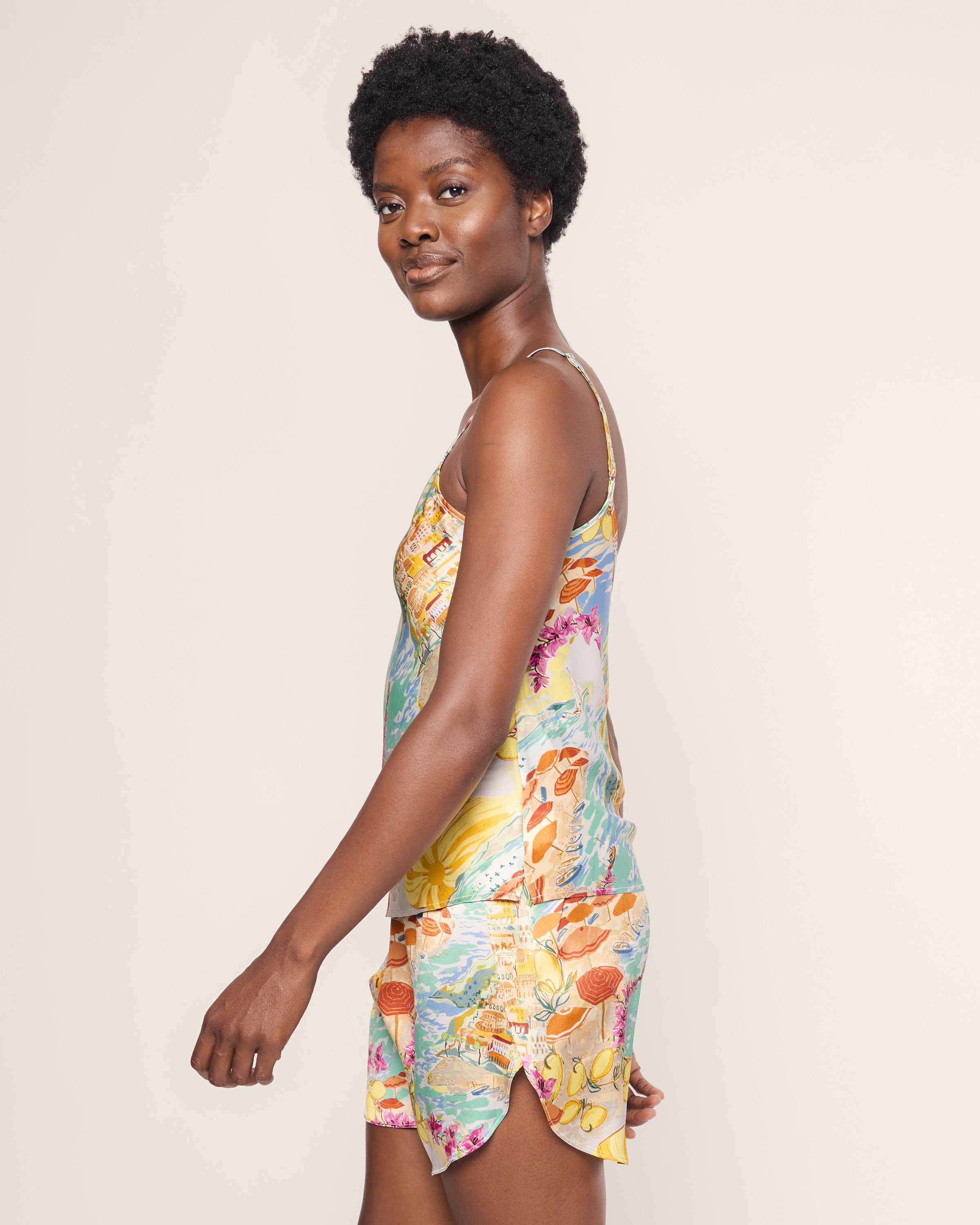 A person with short curly hair smiles, standing sideways and wearing Petite Plumes Womens Silk Cami Short Set in La Dolce Vita. The plain, light-colored background accentuates the vibrant floral pattern of the luxurious ensemble.