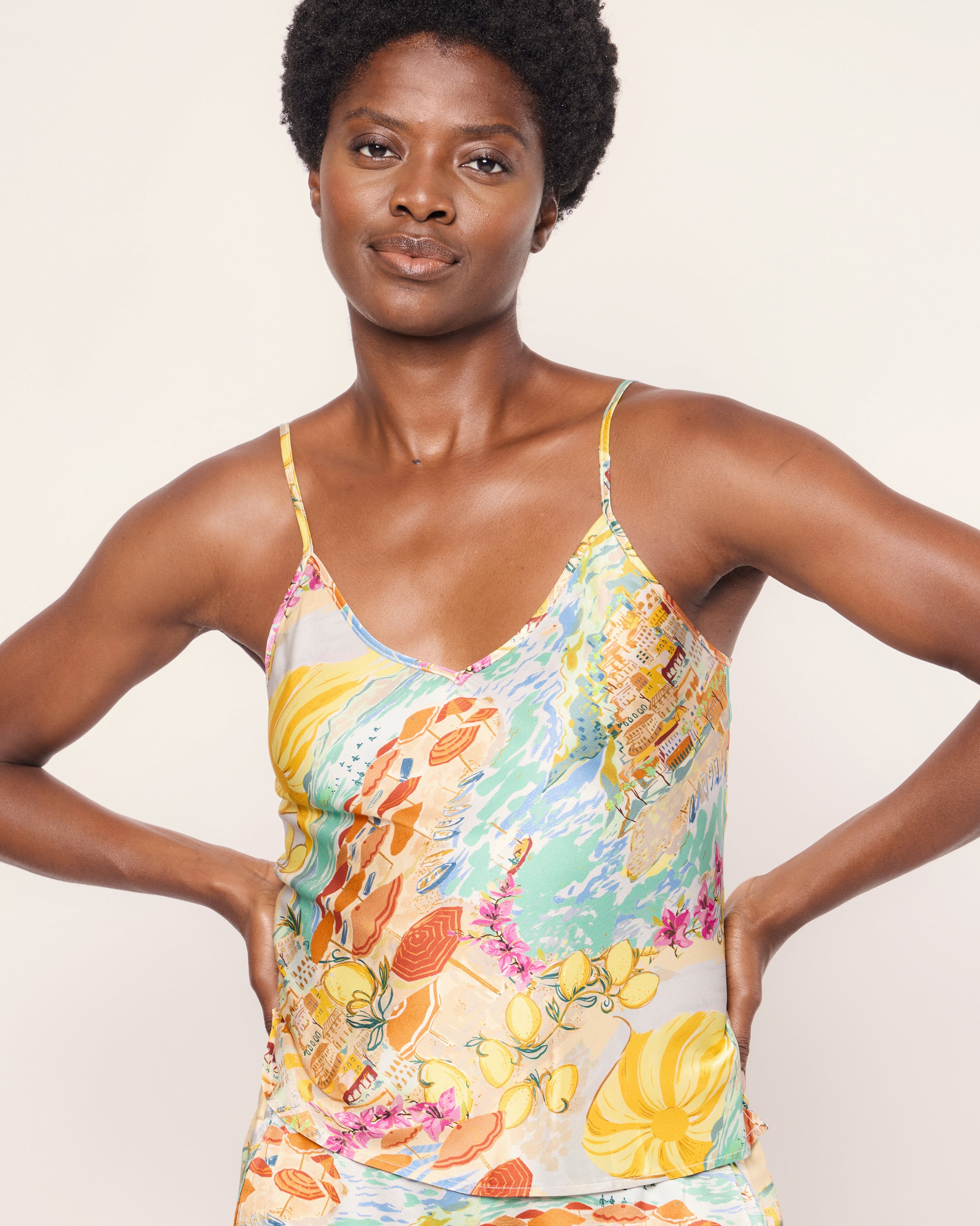 A person is wearing the Womens Silk Cami Short Set in La Dolce Vita by Petite Plume, featuring a colorful pattern with floral and abstract elements in shades of yellow, orange, blue, and pink. They stand with hands on hips against a plain white background.