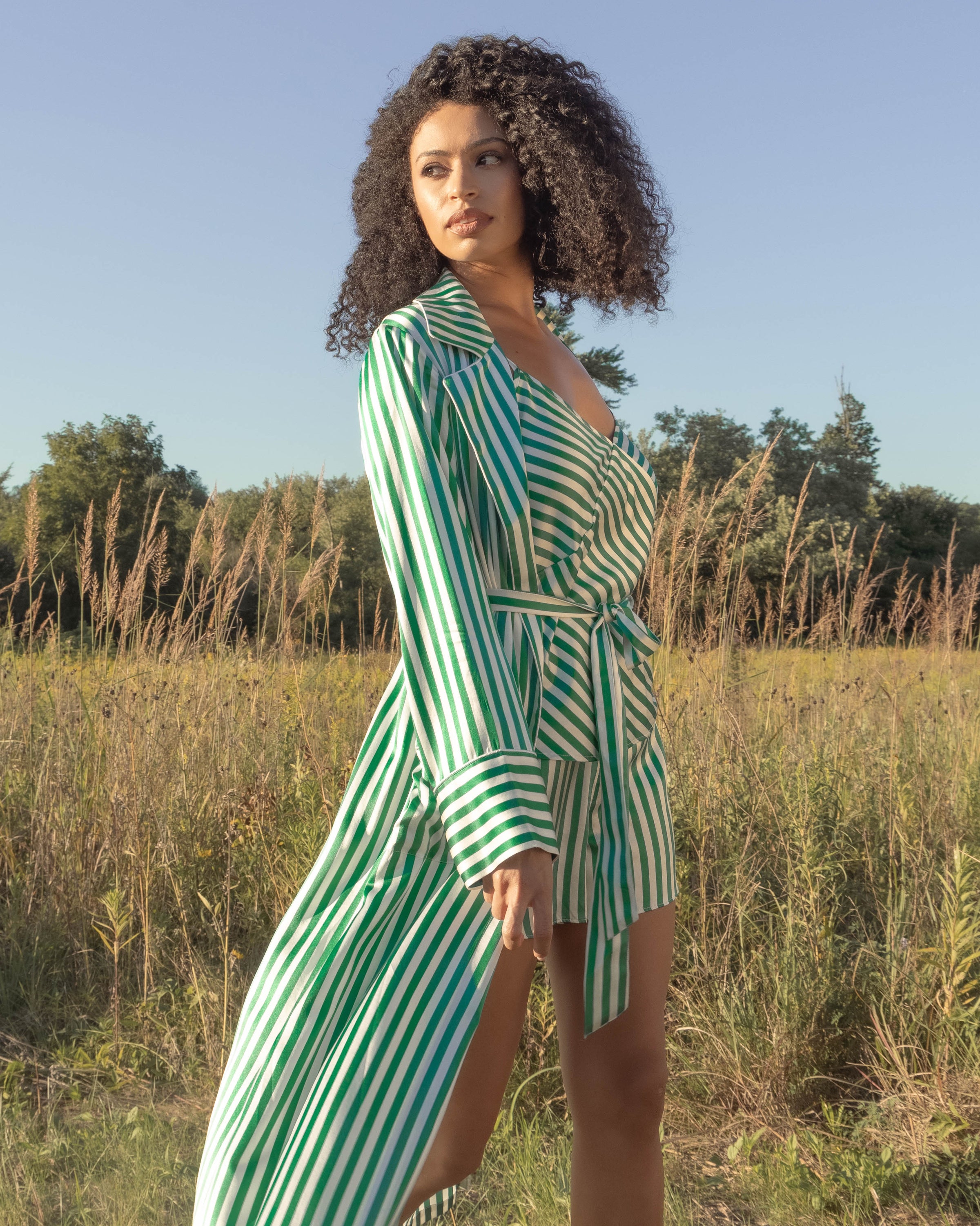 In a field, a woman wears the Womens Silk Cami Short Set in Green Stripe by Petite Plume. Her curly hair cascades around her shoulders as she gazes aside, amidst tall grass and trees under a clear blue sky, evoking the elegance of mulberry silk luxury sleepwear.