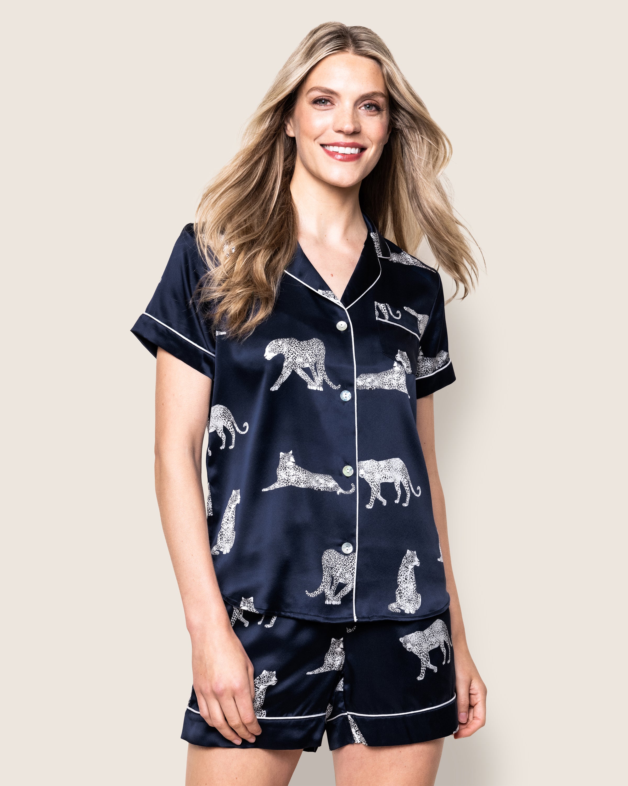 A woman with long blonde hair smiles in luxurious sleepwear by Petite Plume, clad in a navy silk pajama set called the Panthère de Nuit. This stylish set features short sleeves and shorts, adorned with white cheetah and zebra designs, plus a chic button-up top.