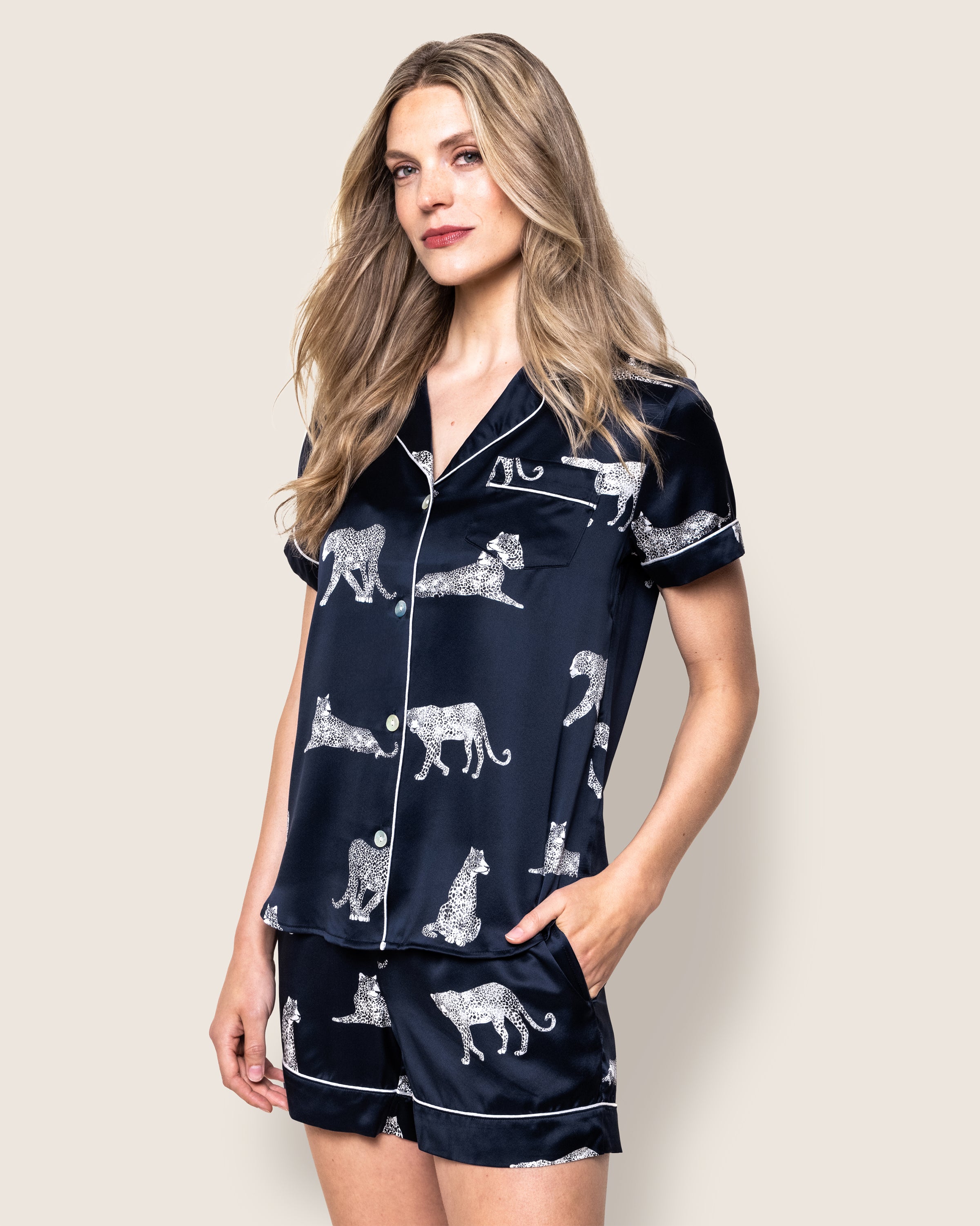 A woman in Petite Plumes Womens Silk Pajama Short Set in Panthère de Nuit with white animal prints poses against a plain background. Her luxurious sleepwear complements her long, wavy blonde hair as she casually rests her hands in her pockets.