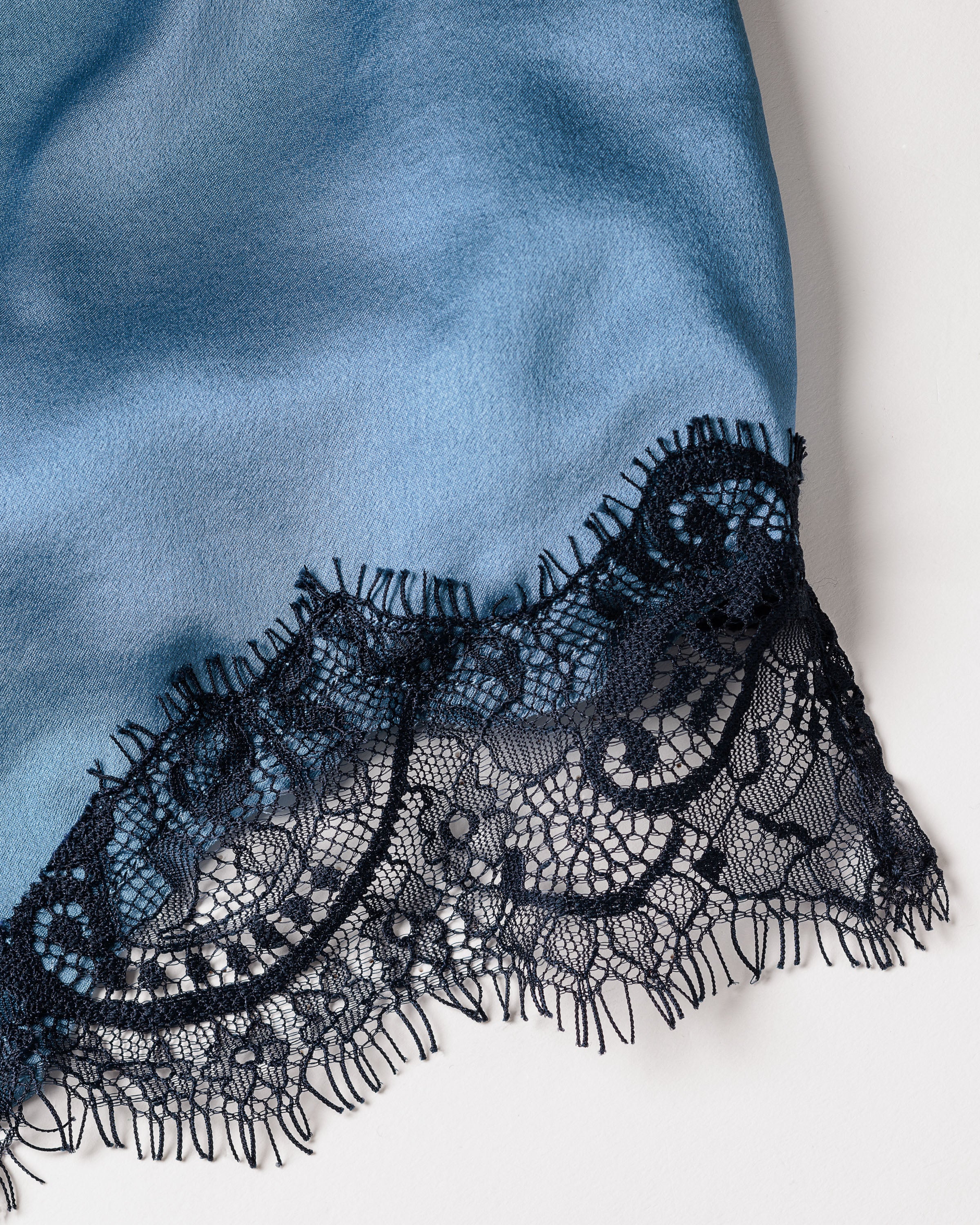 A close-up of the Womens Silk Lace Cami Short Set in Mystique Blue by Petite Plume, showcasing smooth mulberry silk with a decorative black floral lace trim. The intricate detailing makes it ideal for luxurious sleepwear.