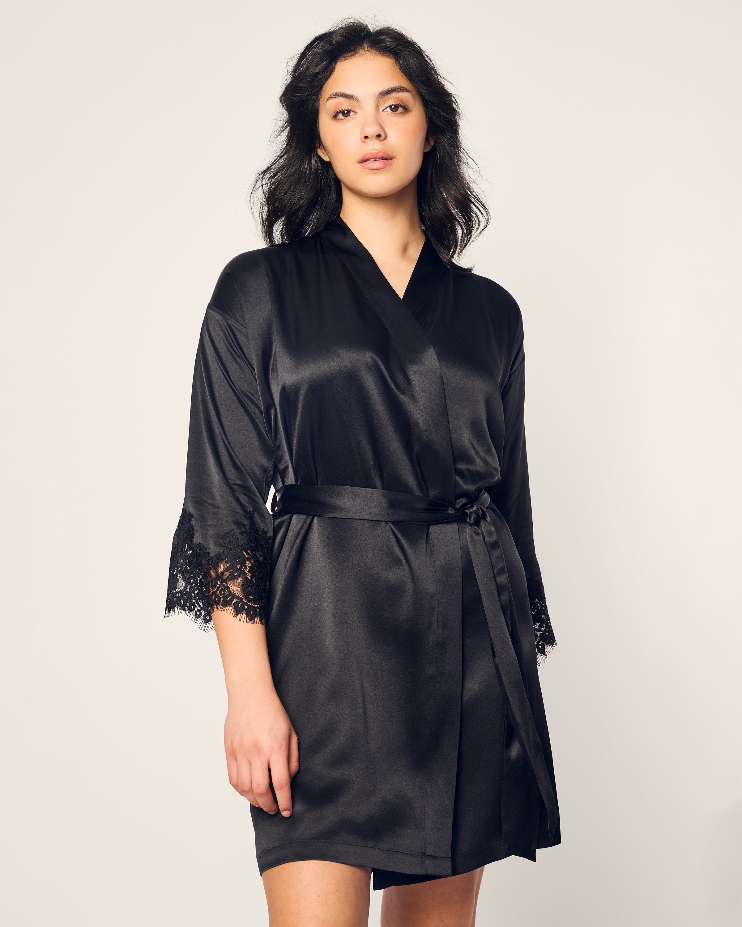 The individual dons a Womens Silk Lace Robe in Black by Petite Plume, crafted from luxurious mulberry silk with lace-trimmed sleeves. Standing against a light background, they gaze at the camera. The belted waist enhances elegance and comfort, akin to fine silk sleepwear.