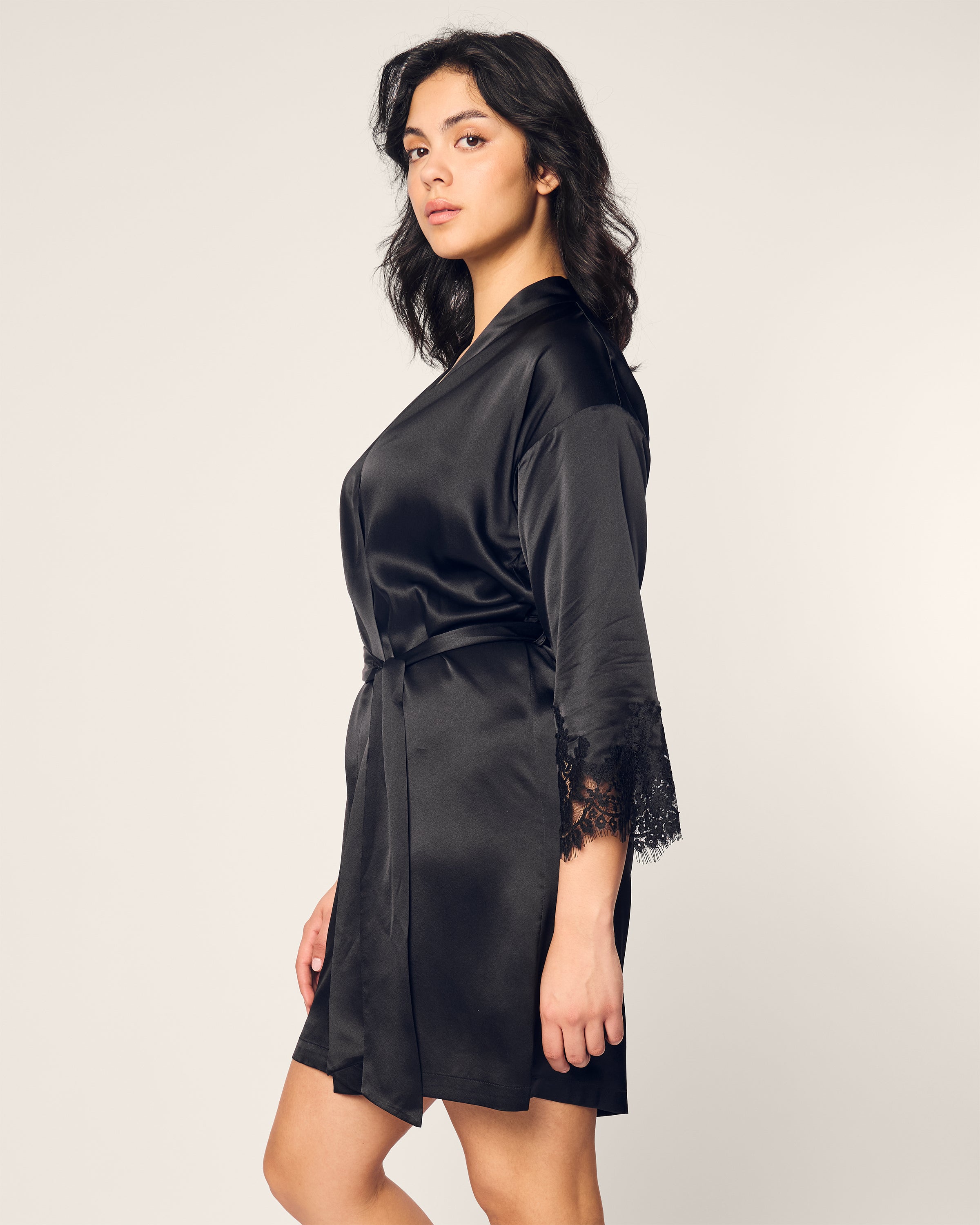 A person with shoulder-length dark hair stands sideways, looking at the camera, wearing a Petite Plume Womens Silk Lace Robe in Black. The robe is crafted from mulberry silk with lace sleeve details against a plain off-white background.
