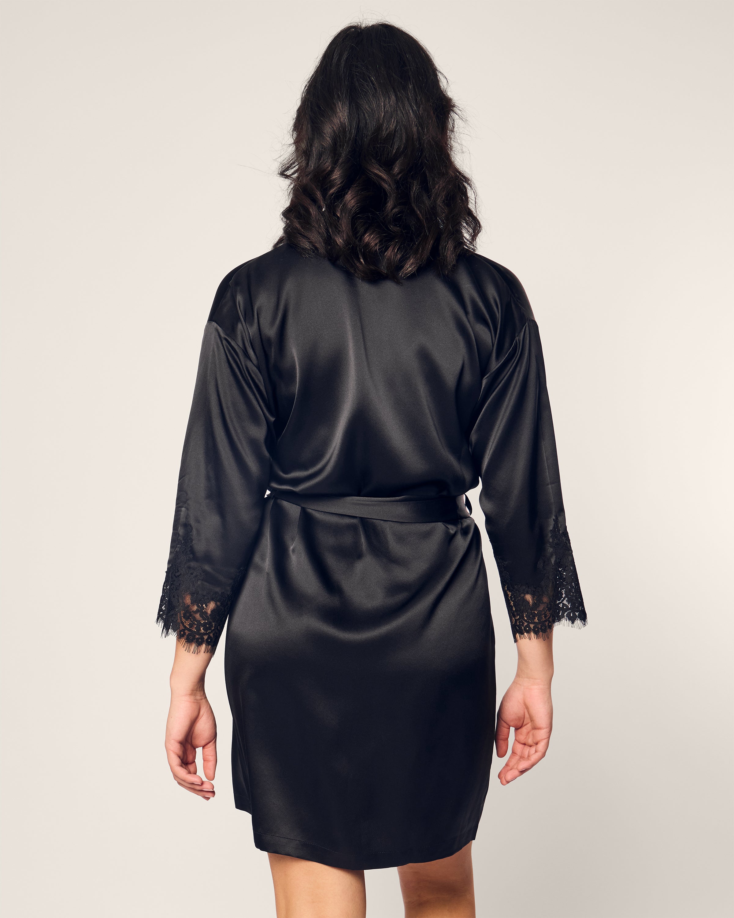 Someone with shoulder-length hair stands with their back to the camera, wearing a luxurious Womens Silk Lace Robe in Black by Petite Plume. The mulberry silk sleepwear adds elegance against the plain, light-colored background.