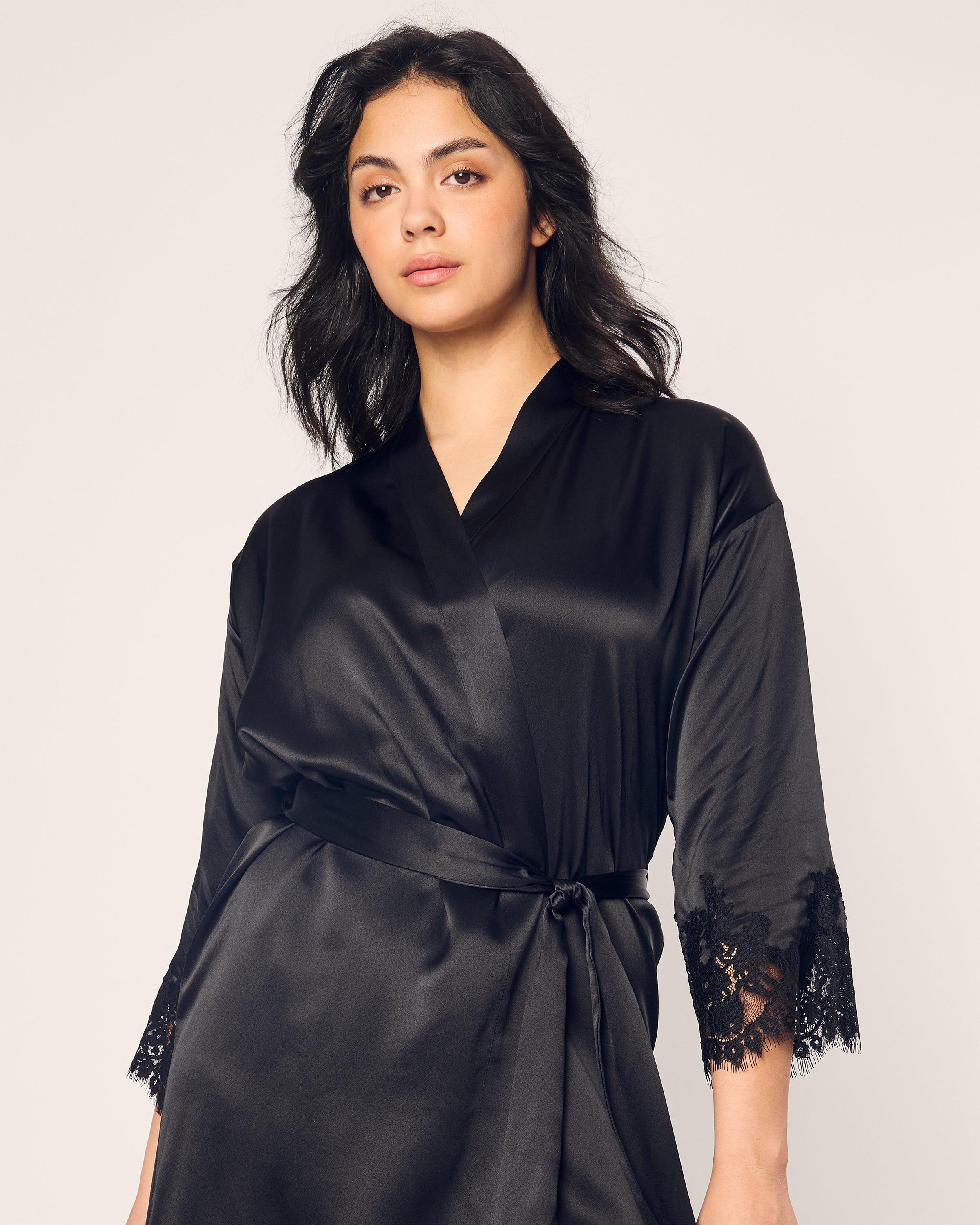 A person with long dark hair poses confidently, wearing the Petite Plume Womens Silk Lace Robe in Black, featuring lace-trimmed sleeves. Luxuriously made from mulberry silk, the robe is tied at the waist against a plain light background.