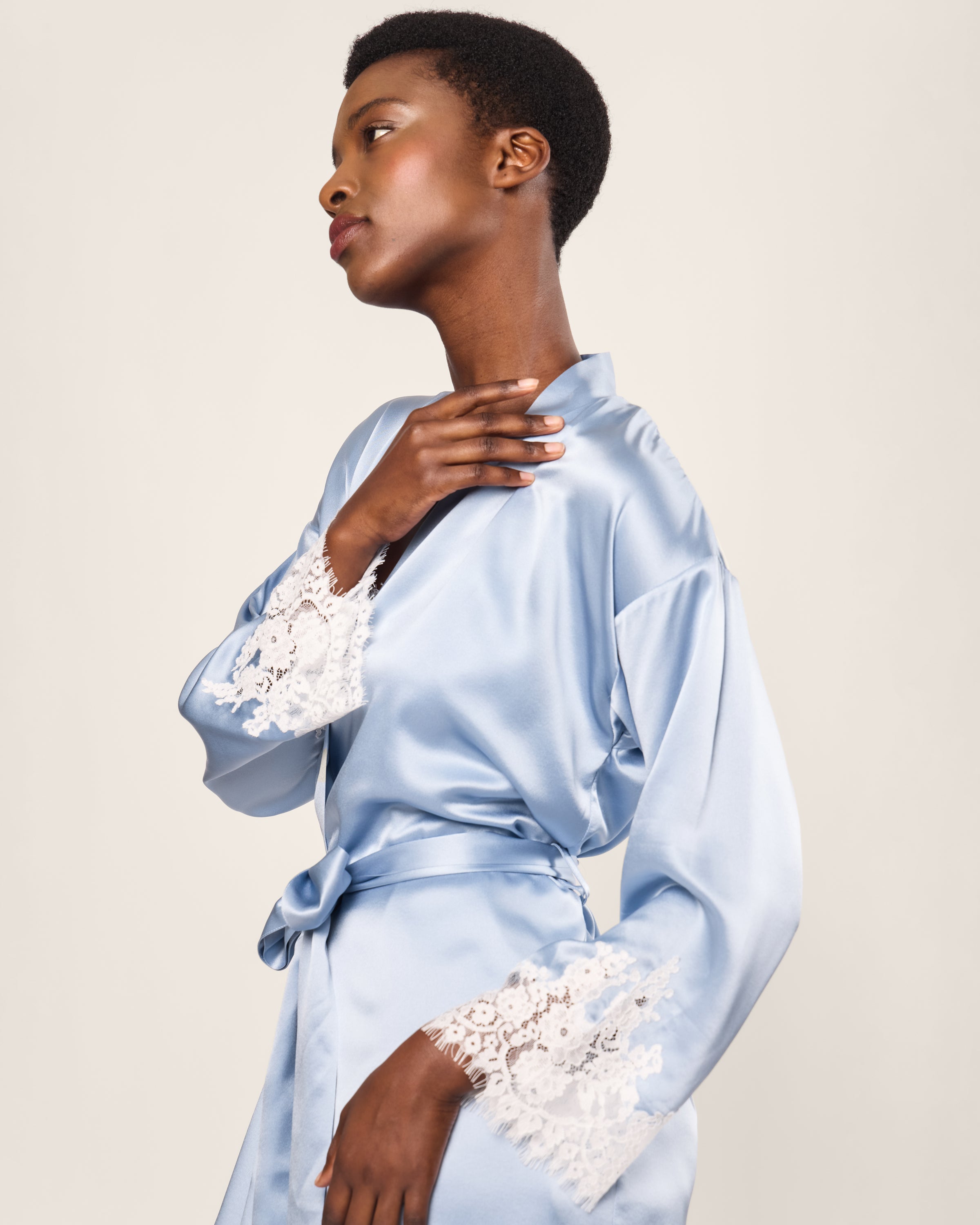 A person poses against a plain background in the Womens Silk Lace Robe in Periwinkle by Petite Plume. The robe, featuring lace cuffs and a tied waist, is gently touched at the collar by one hand, as the individual gazes slightly upward, evoking luxurious elegance.