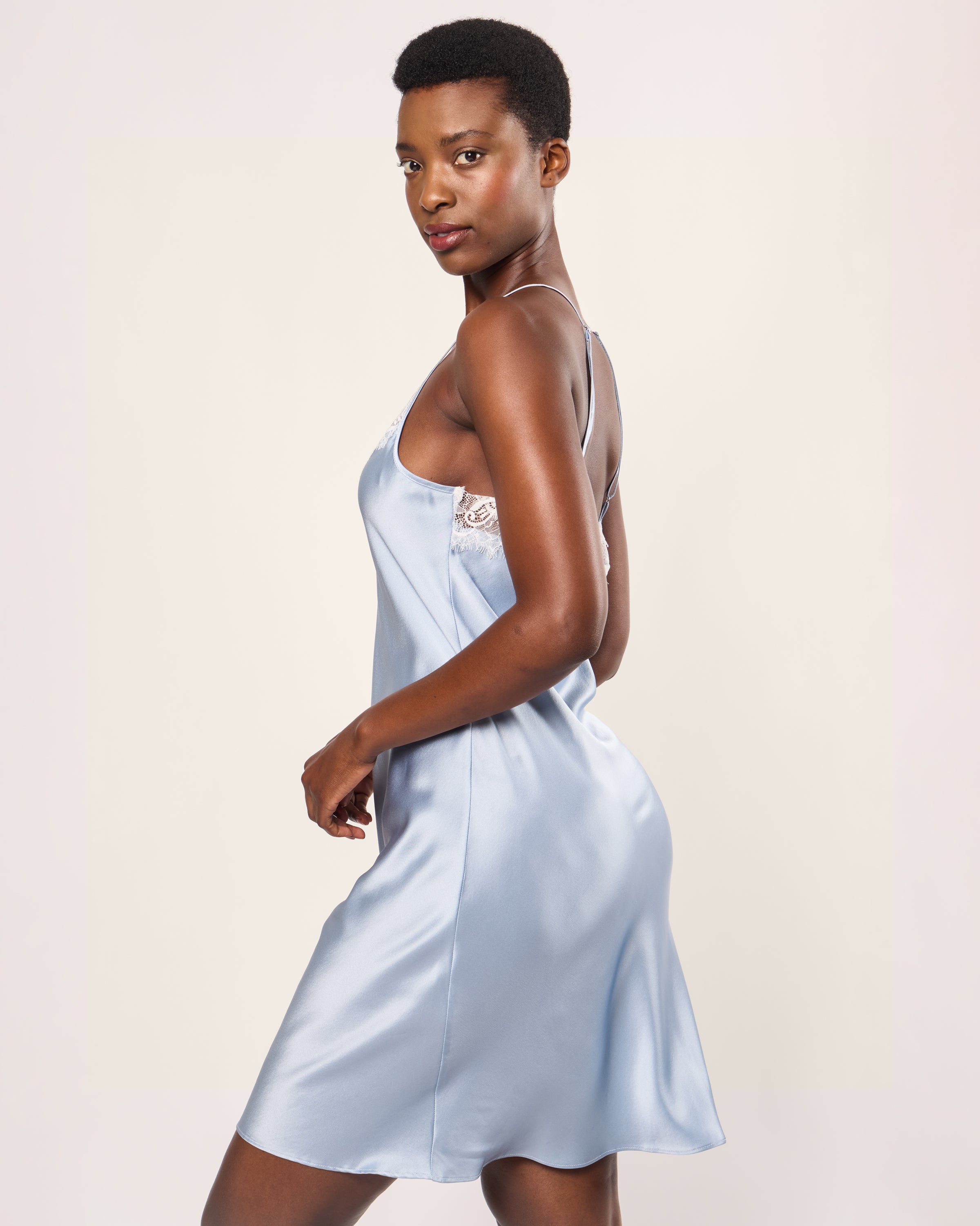 On a plain backdrop, a model showcases the Womens Silk Lace Slip Dress in Periwinkle by Petite Plume. This piece reflects minimalist nightdress design with its delicate lace accents, spaghetti straps, and knee-length hem, embodying luxury sleepwear through understated elegance and a chic silhouette.