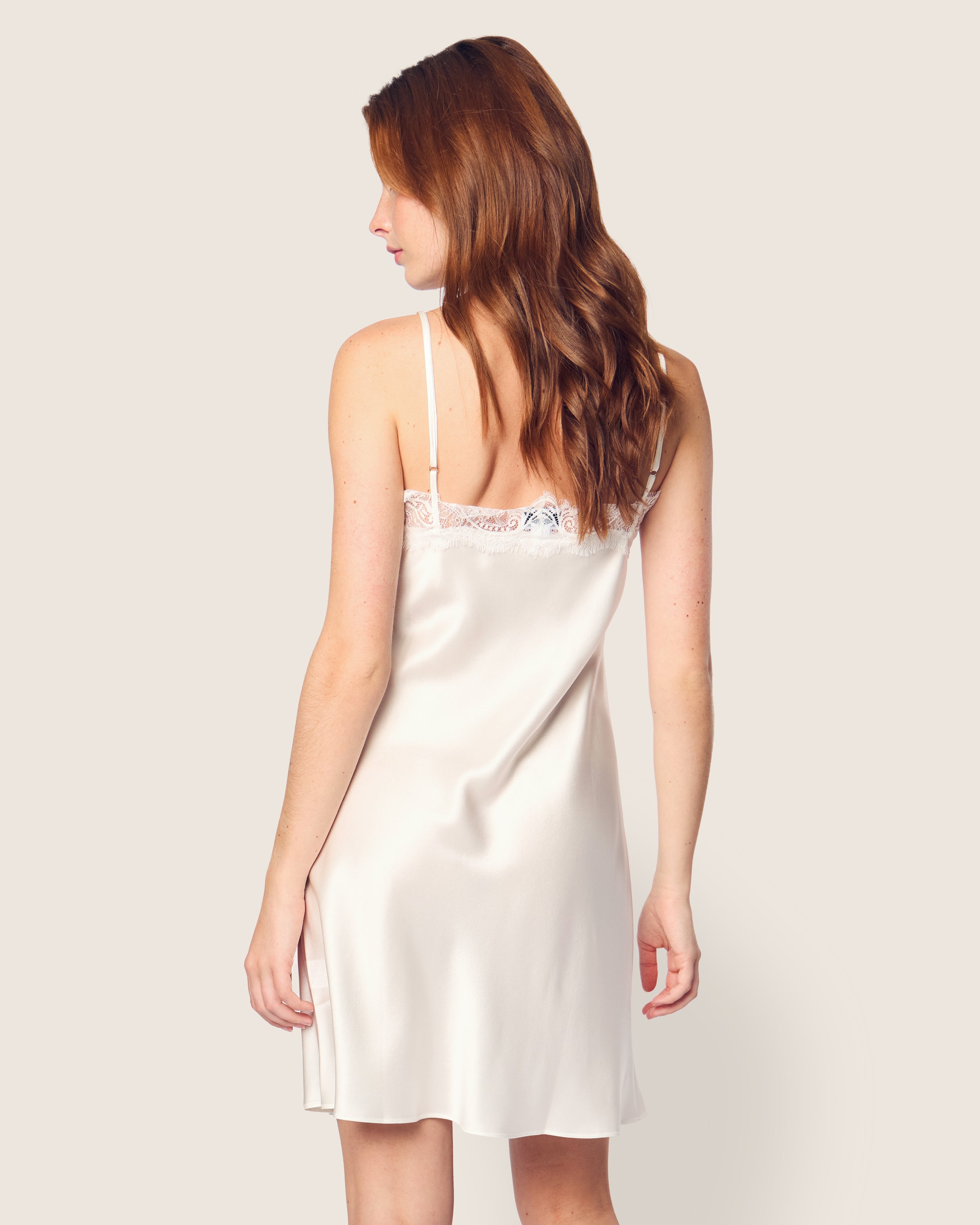 A woman with long brown hair embodies minimalist fantasy in a luxurious Womens Silk Lace Slip Dress in White by Petite Plume, gracefully facing away from the camera against an off-white background.