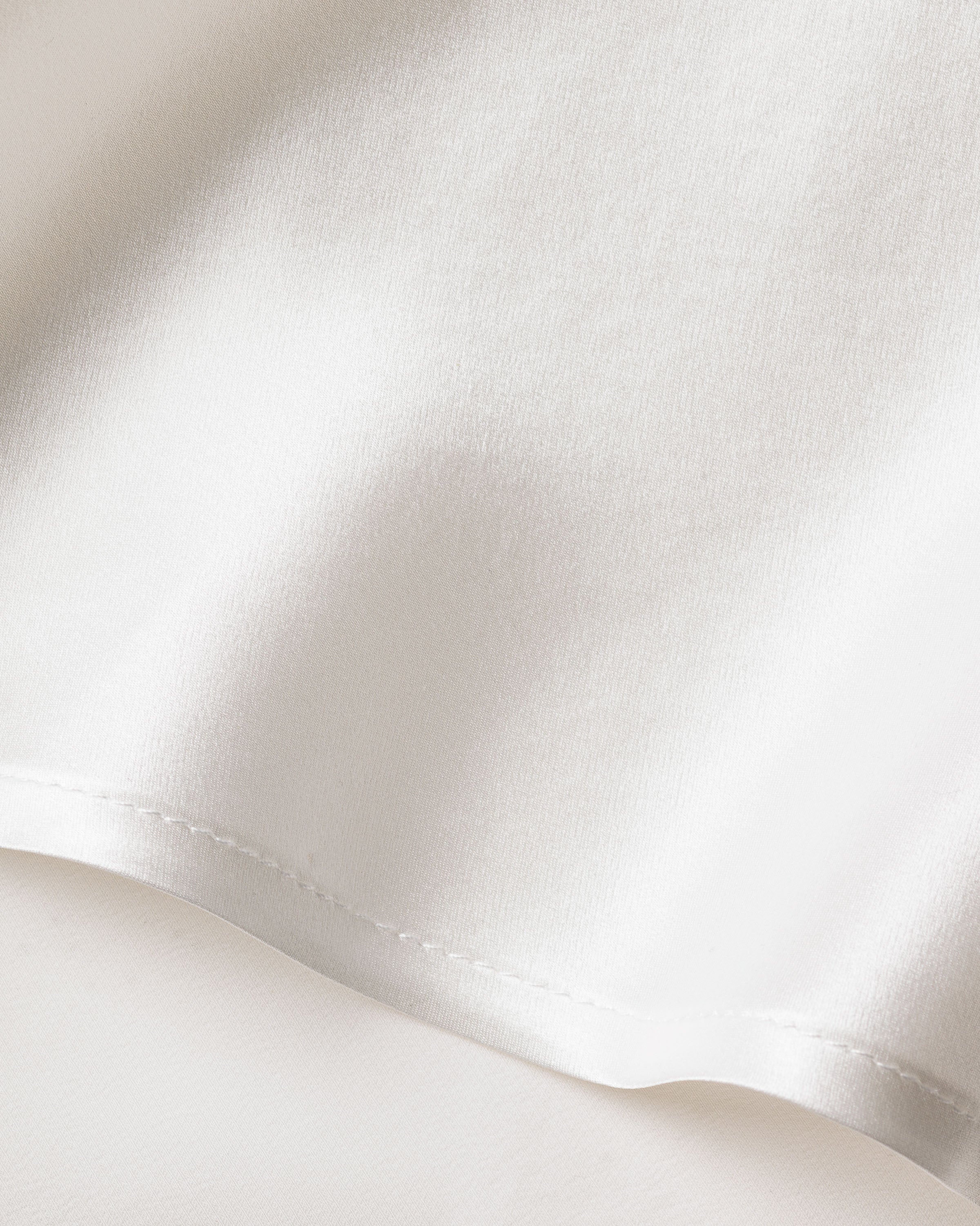 A close-up of a smooth, silk fabric from the Womens Silk Lace Slip Dress in White by Petite Plume reveals a luxurious sleepwear sheen. The material gently folds with a soft texture, capturing light and casting slight shadows.
