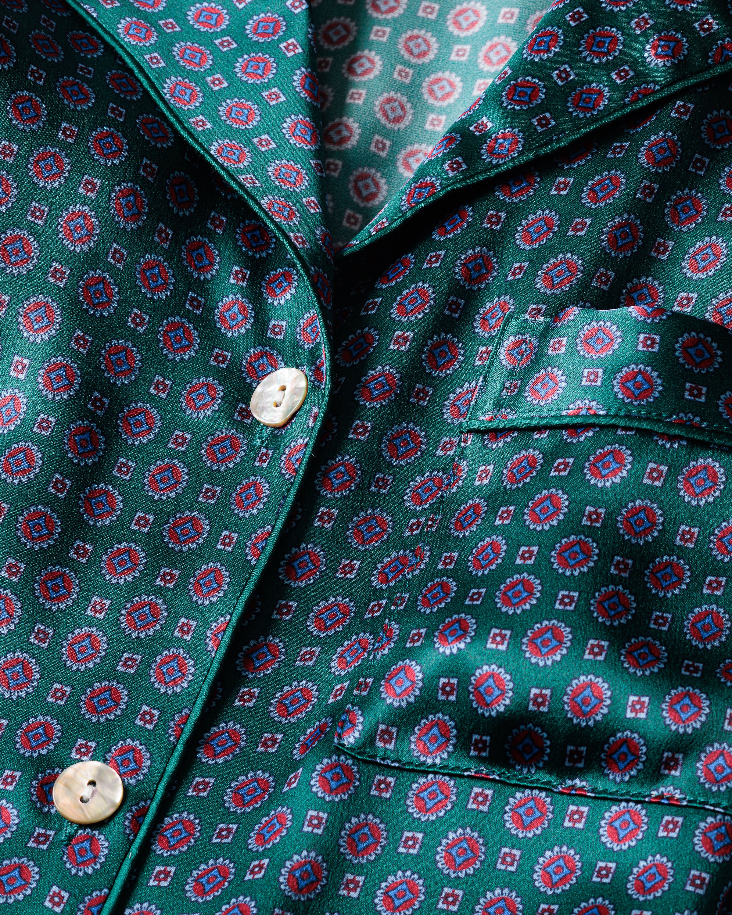 A close-up of Petite Plumes Womens Silk Long Sleeve Short Set in Emerald Foulard showcases red and blue geometric shapes. With a pocket and shiny white buttons, it exudes luxurious, timeless pajama-style elegance.