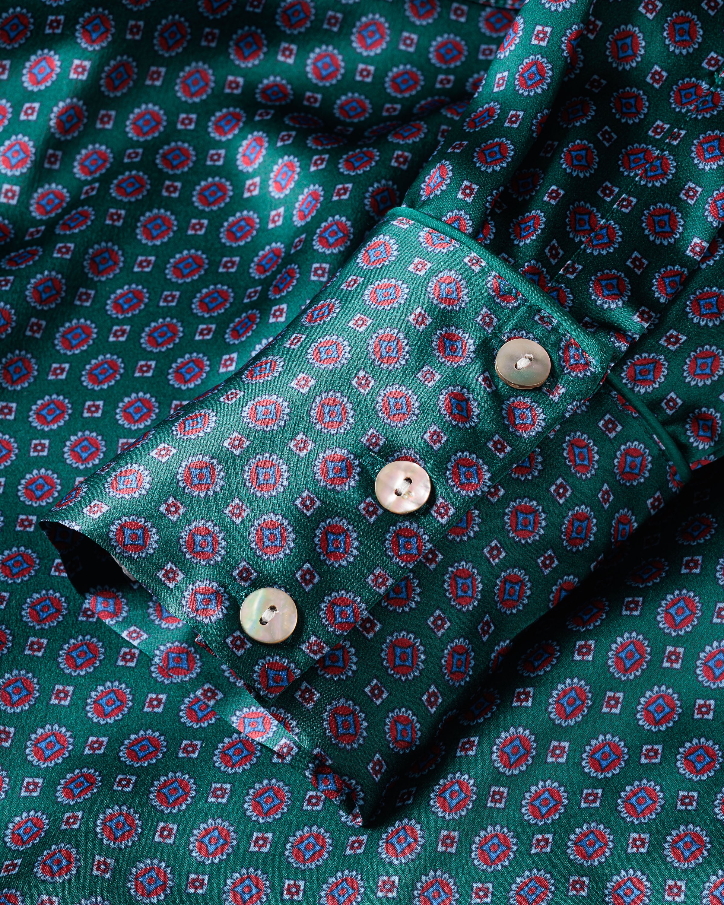 Close-up of an emerald foulard fabric with a red and blue geometric pattern, showcasing a shirt cuff with four white buttons. Crafted from Mulberry silk, this luxurious textile by Petite Plume embodies the elegance and comfort of the Womens Silk Long Sleeve Short Set in timeless pajama design.