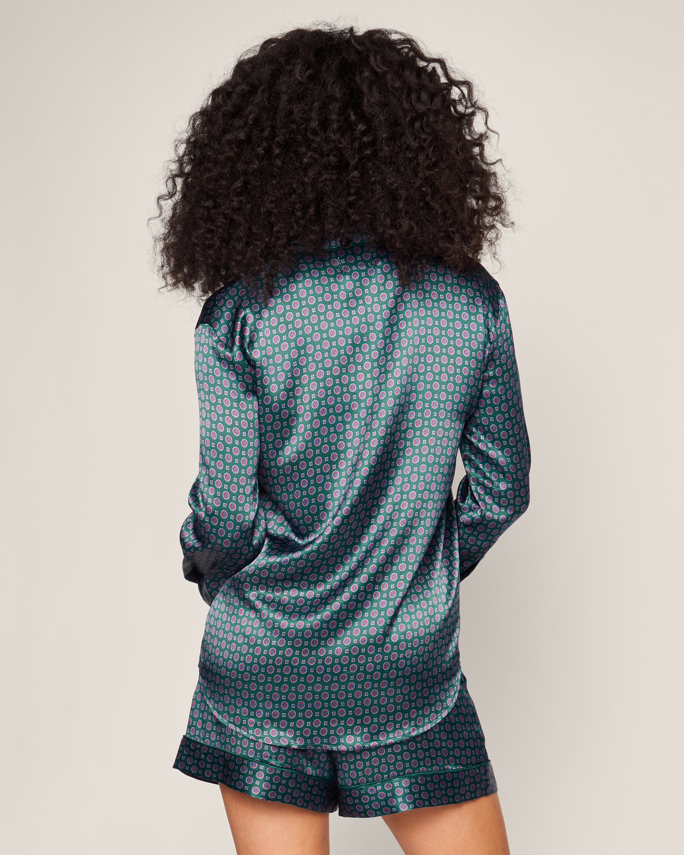 A person with curly hair is seen from behind, wearing Petite Plumes Womens Silk Long Sleeve Short Set in Emerald Foulard. The luxurious Mulberry silk features a teal and pink geometric pattern. A plain beige background highlights the timeless sleepwear with above-the-knee shorts.