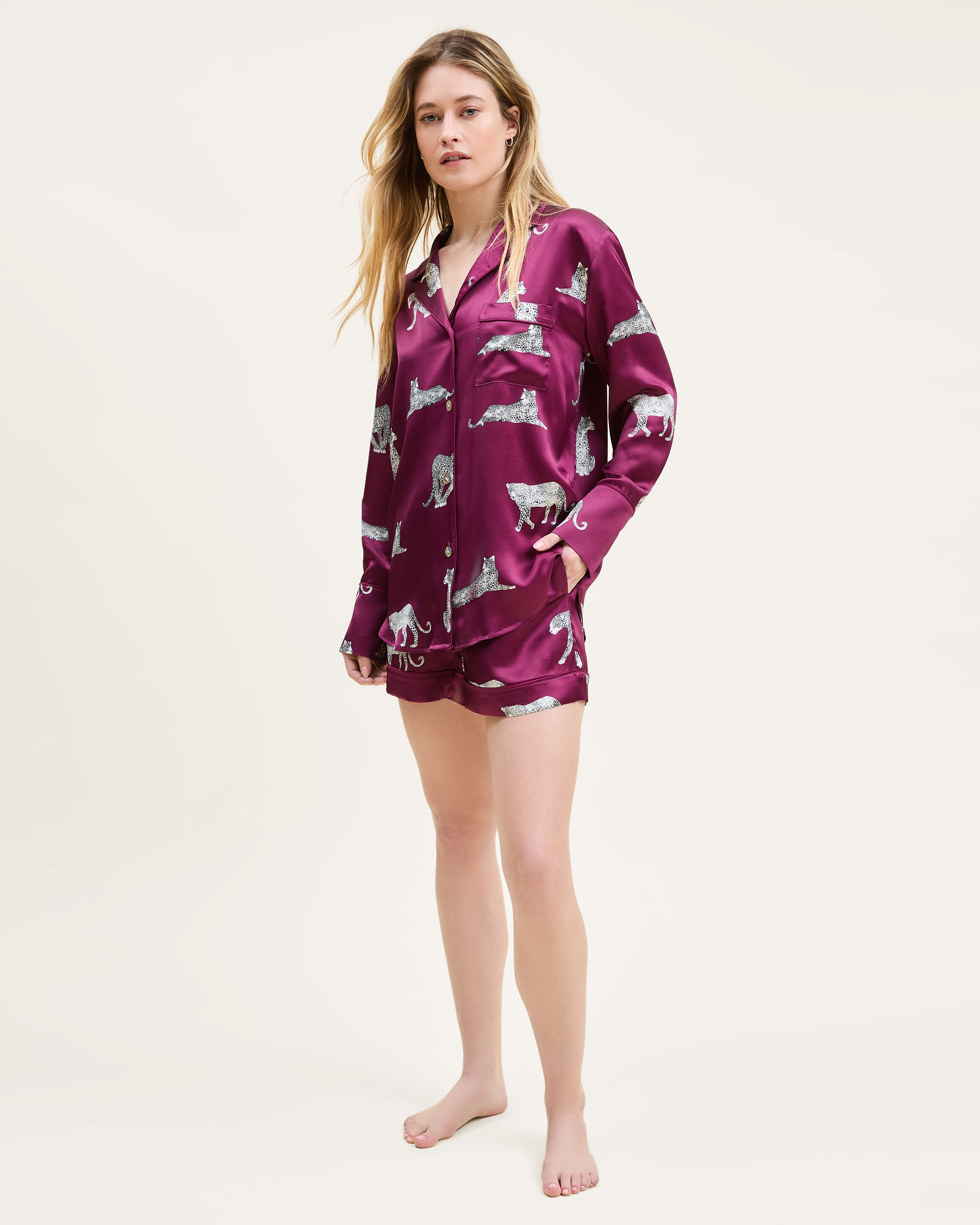 The Women's Silk Long Sleeve Short Set in Merlot Panthère features a dark red satin pajama set with a button-up top and shorts, adorned with white illustrated leopards in various poses.