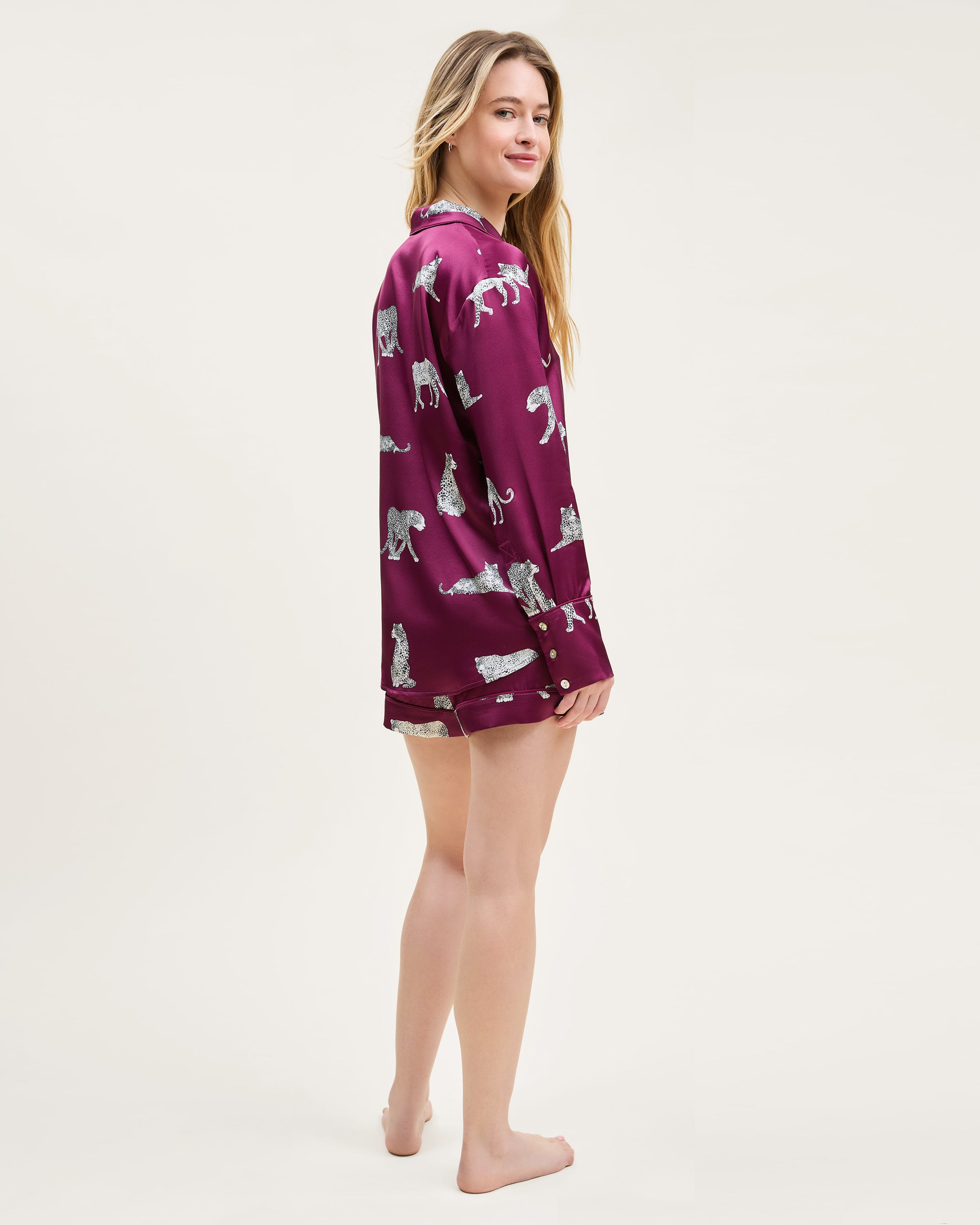 The Women's Silk Long Sleeve Short Set in Merlot Panthère features a dark red satin pajama set with a button-up top and shorts, adorned with white illustrated leopards in various poses.