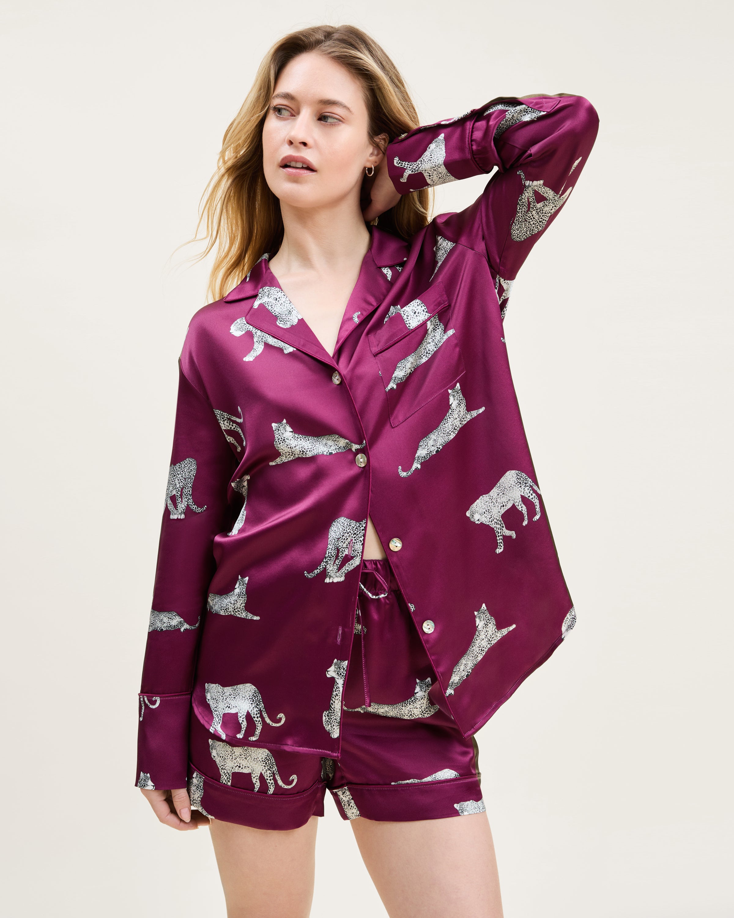 The Women's Silk Long Sleeve Short Set in Merlot Panthère features a dark red satin pajama set with a button-up top and shorts, adorned with white illustrated leopards in various poses.