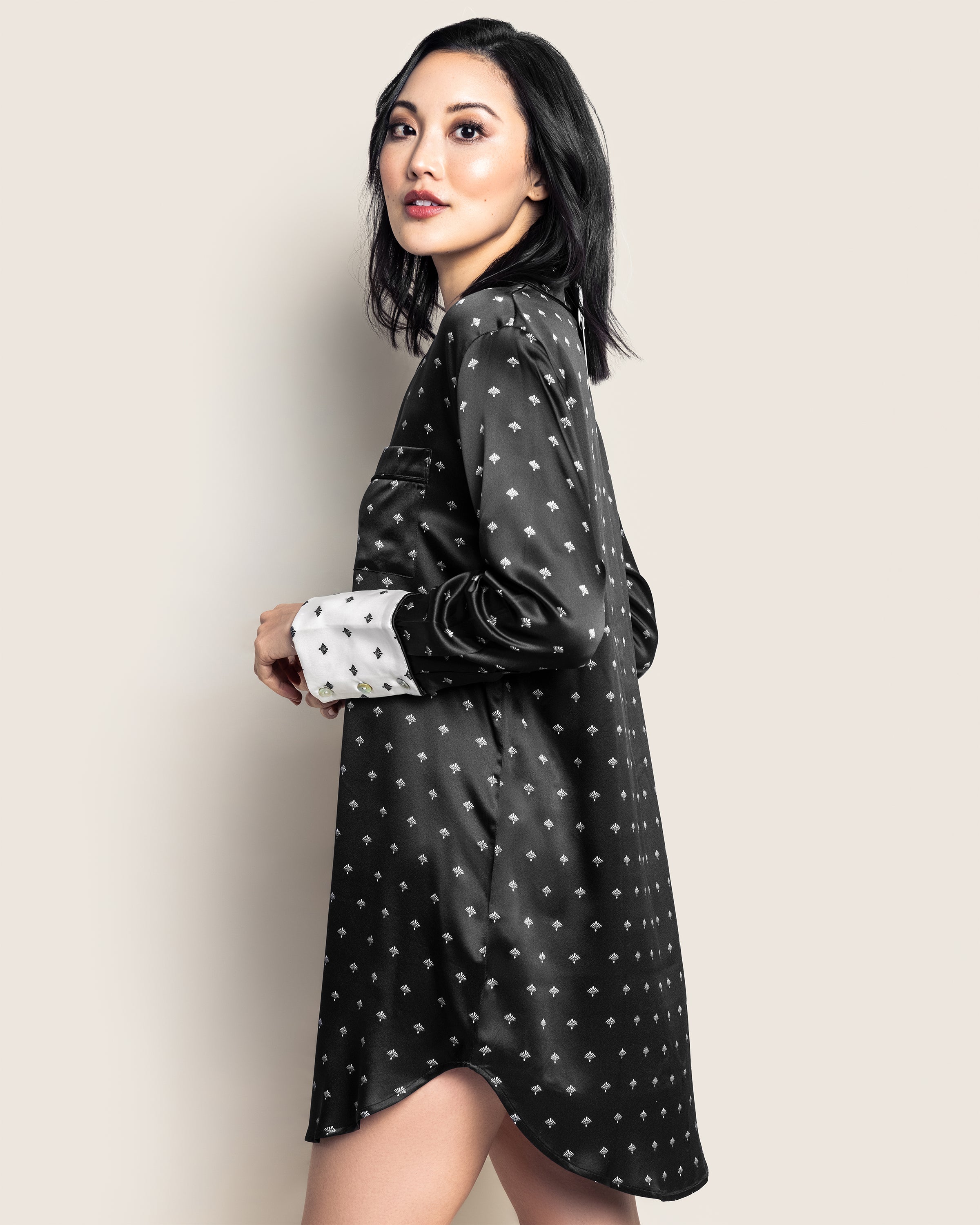 A woman with shoulder-length black hair wears a Petite Plume Womens Silk Nightshirt in Black Art Nouveau, featuring white cuffs. She stands against a plain beige background, gazing slightly right with an enigmatic air reminiscent of Truman Capotes muses.