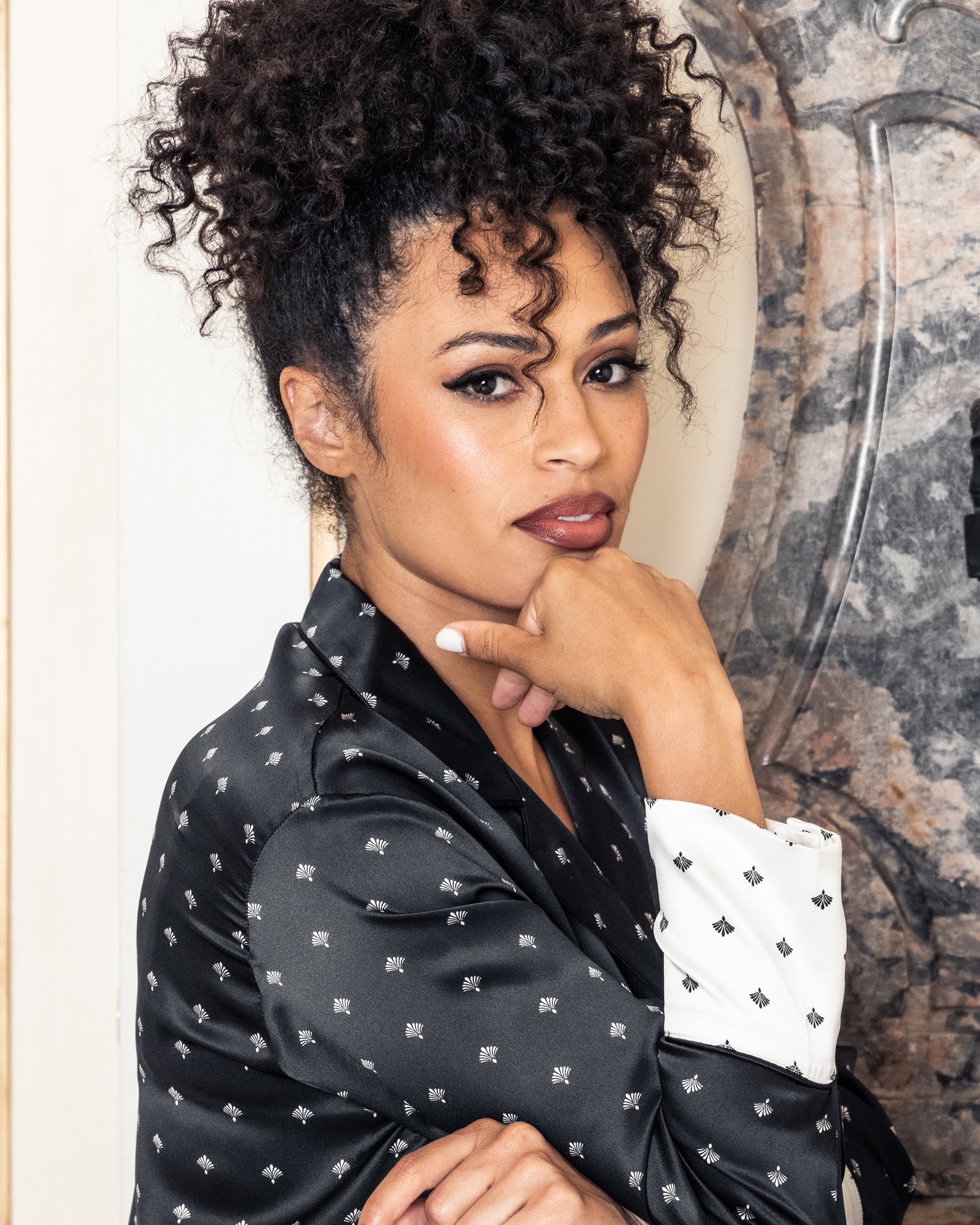 A curly-haired woman poses thoughtfully, wearing the Petite Plume Womens Silk Nightshirt in Black Art Nouveau. She rests her chin on her hand, gazing into the camera against an ornate backdrop that enhances the sophisticated ambiance.